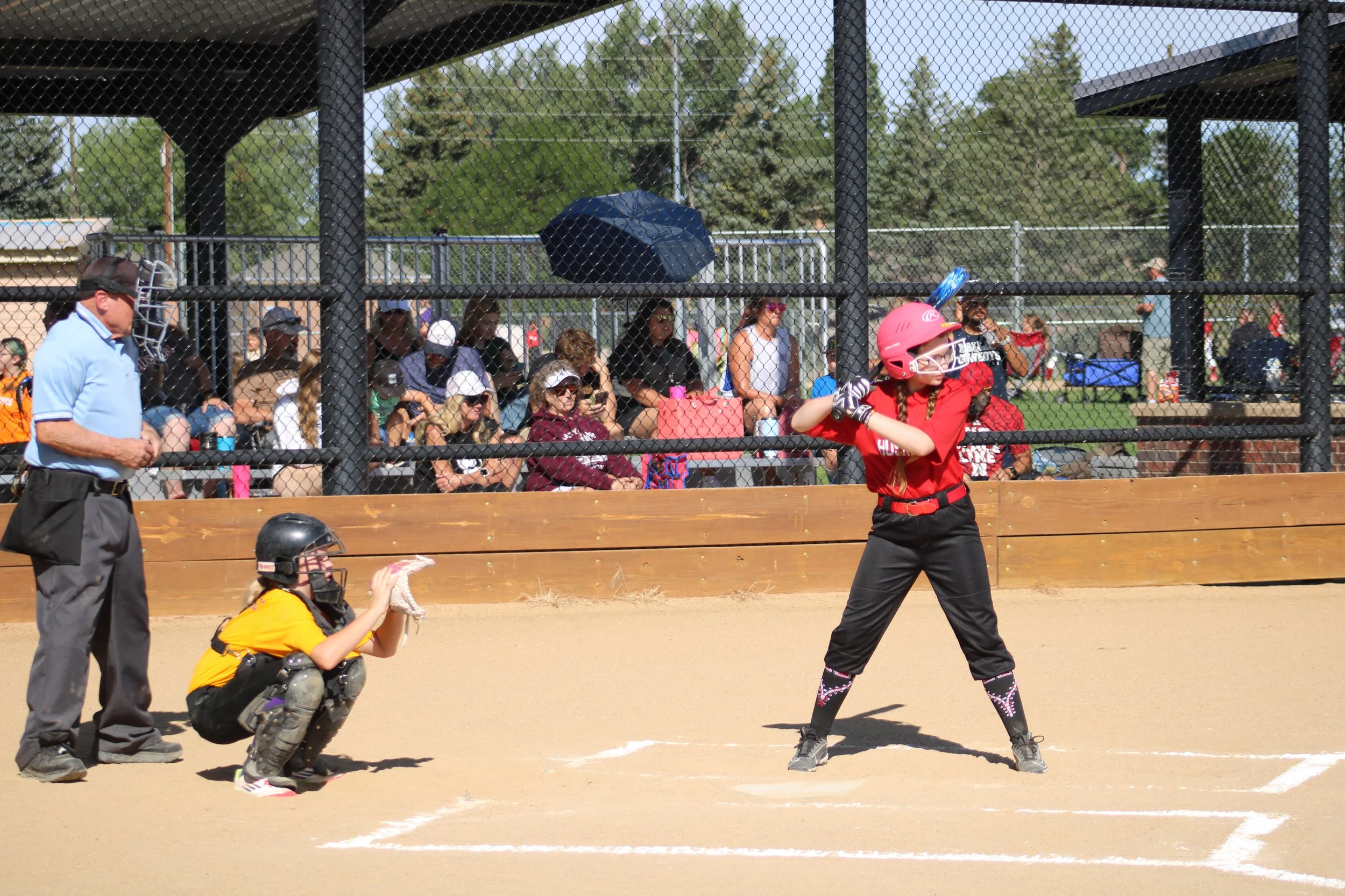 A young girl in red softball uniform and pink helmet batting at a pitched ball at a softball game. A catcher in yellow gear crouches behind home plate, and an umpire in blue shirt stands watching. Spectators seated behind the chain-link fence watch the game.