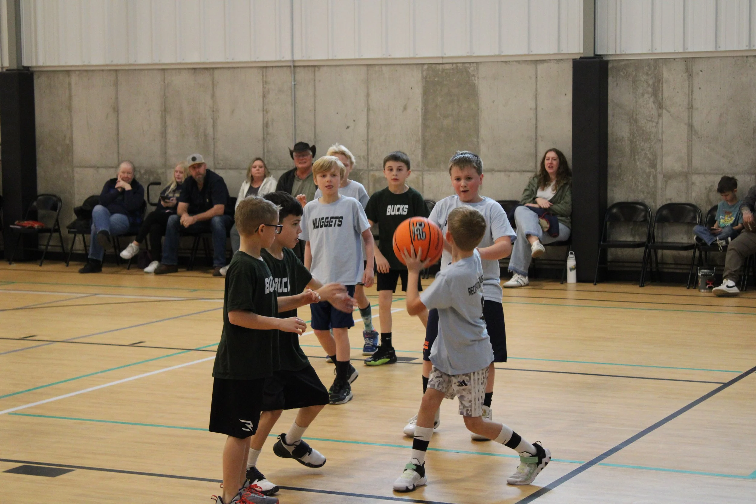 Children playing basketball in an indoor gymnasium, with spectators sitting on chairs along the wall.