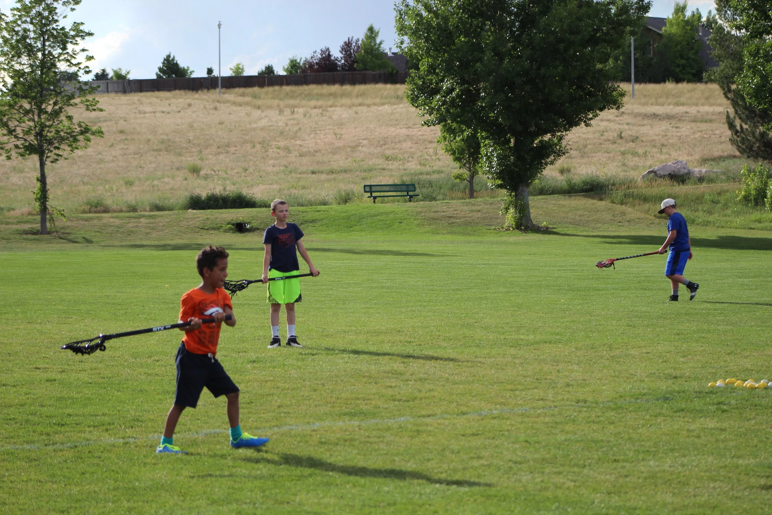 Three boys are practicing golf on a grassy field with trees and a hill in the background.