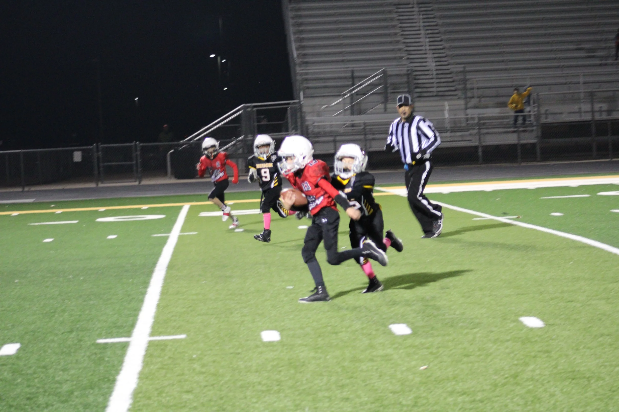 Young kids playing flag football on a brightly lit field at night, with a referee observing in the background.