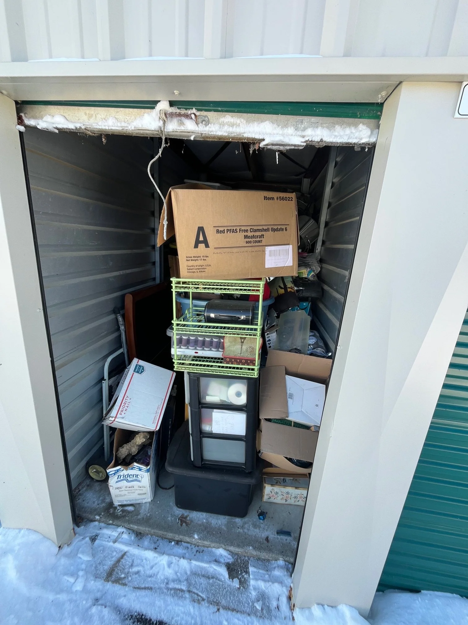 A storage unit filled with miscellaneous boxes, plastic containers, and household items, with snow on the ground outside.