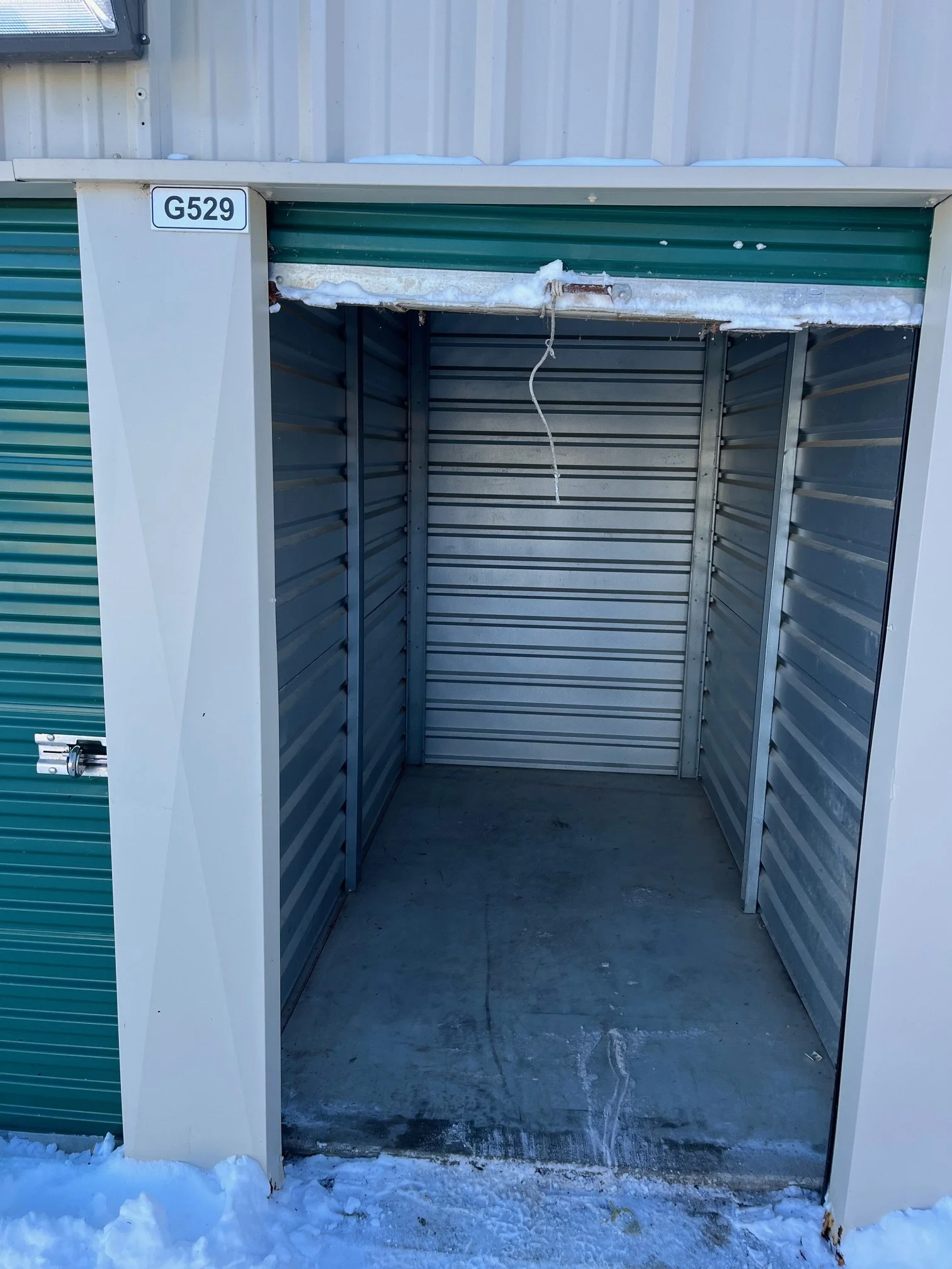 Empty outdoor storage unit with snow on the ground, metal walls, and a partially open roll-up door.