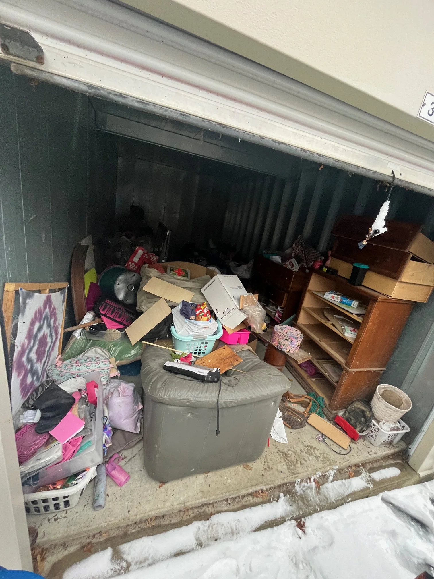 Cluttered garage with various items, including baskets, boxes, a bookshelf, and miscellaneous objects, with snow outside.