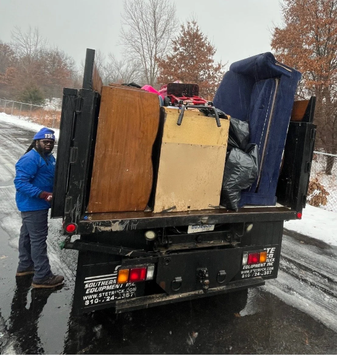 A man in a blue jacket, pants, and a blue hat standing beside a truck filled with furniture and belongings on a snowy, muddy road with leafless trees in the background.