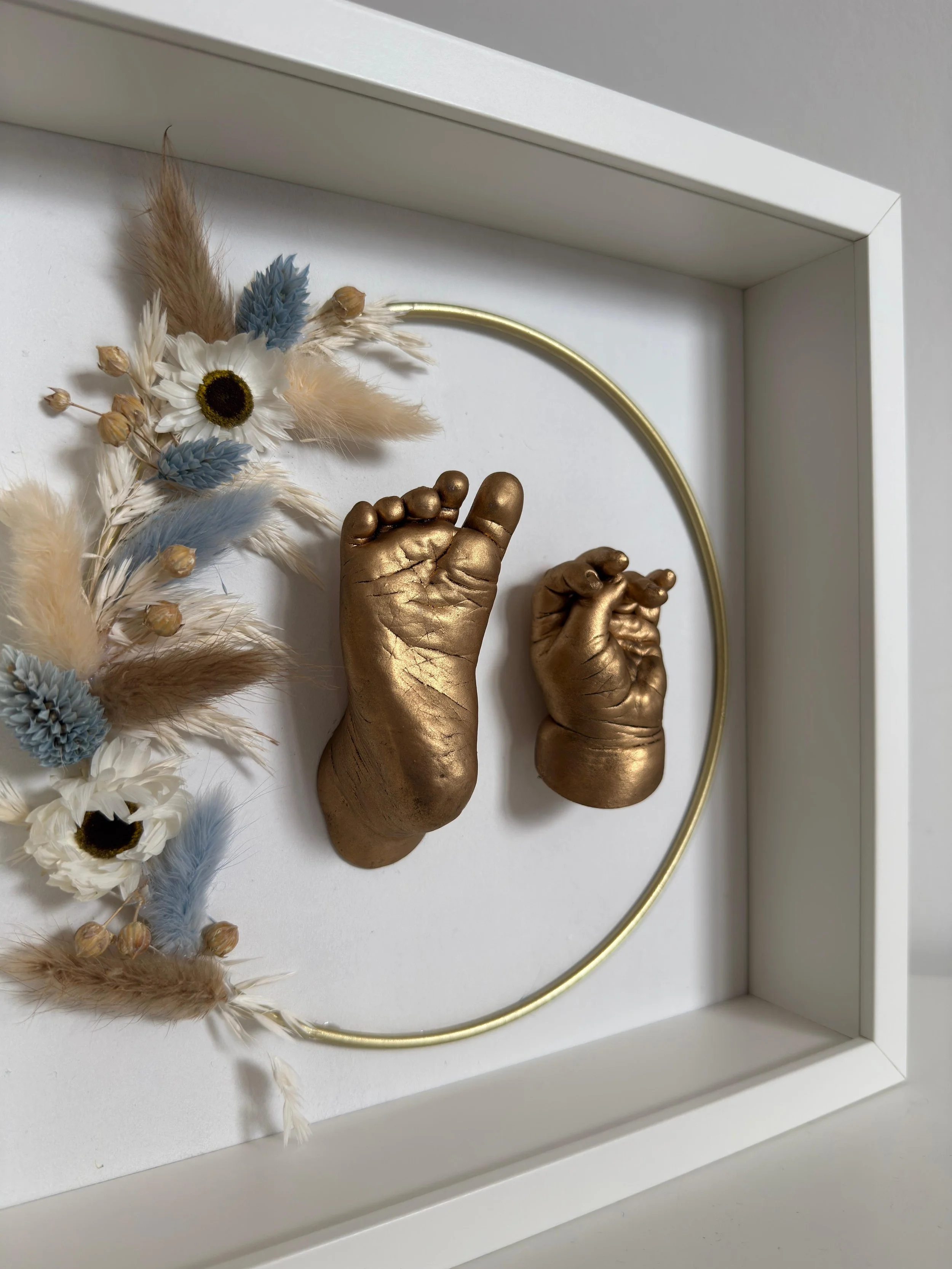 Gouden afdruk van een baby handje en voetje omringd met een gouden ring en droogbloemen