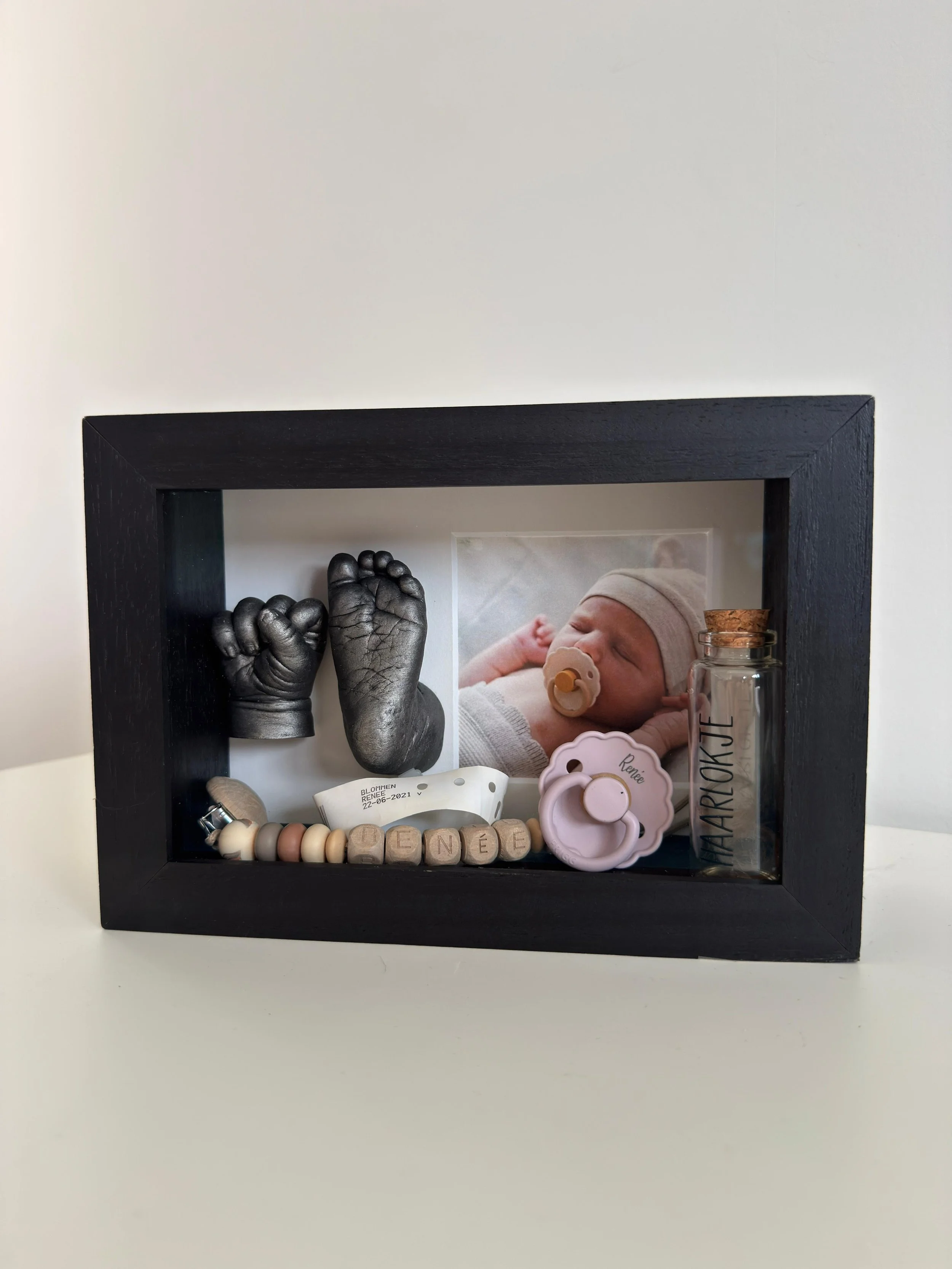 Afdruk van een baby handje en voetje in een diepe 3d kader, samen met een foto van de baby en attributen in de kader.