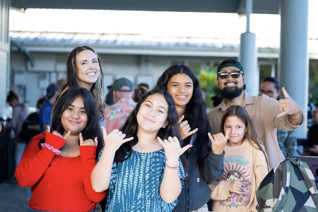 Group of six diverse people smiling and making hand signs, standing outdoors at a social gathering.
