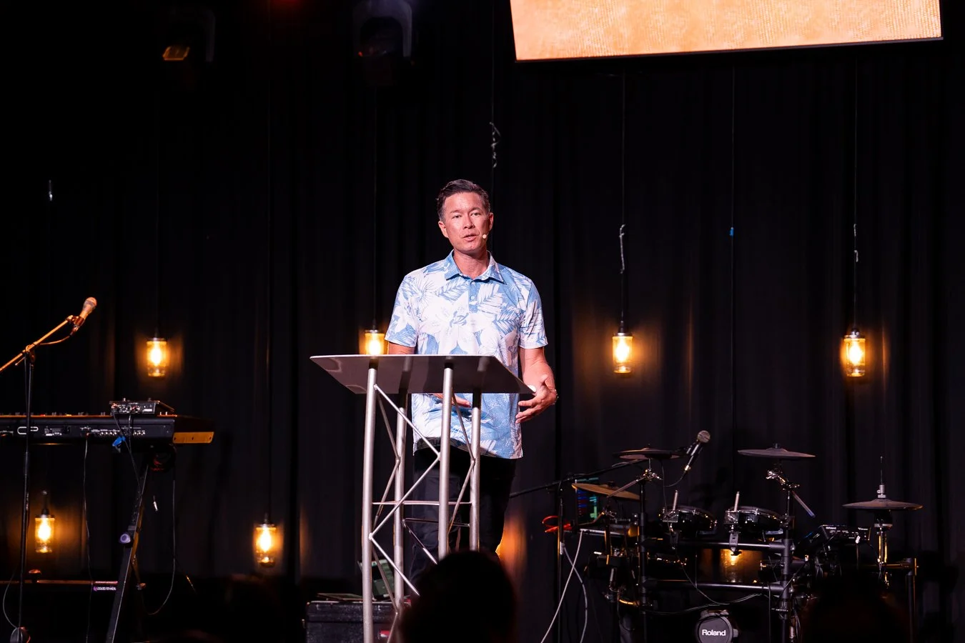 A man standing behind a lectern on a stage with black curtains and hanging Edison-style light bulbs, appears to be giving a presentation or speech, with musical instruments visible to his sides.