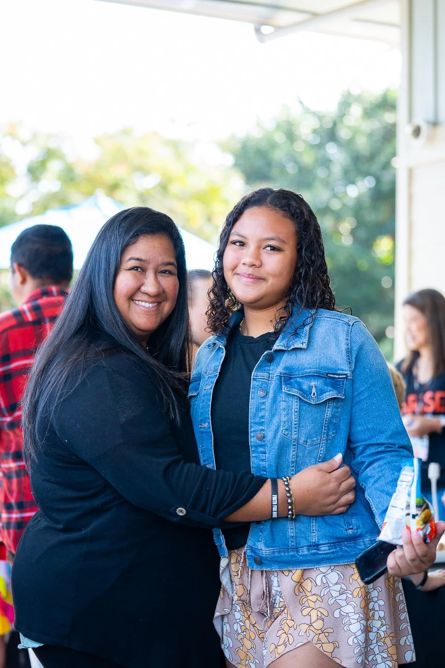 Two women smiling and hugging at an outdoor gathering, with other people in the background.