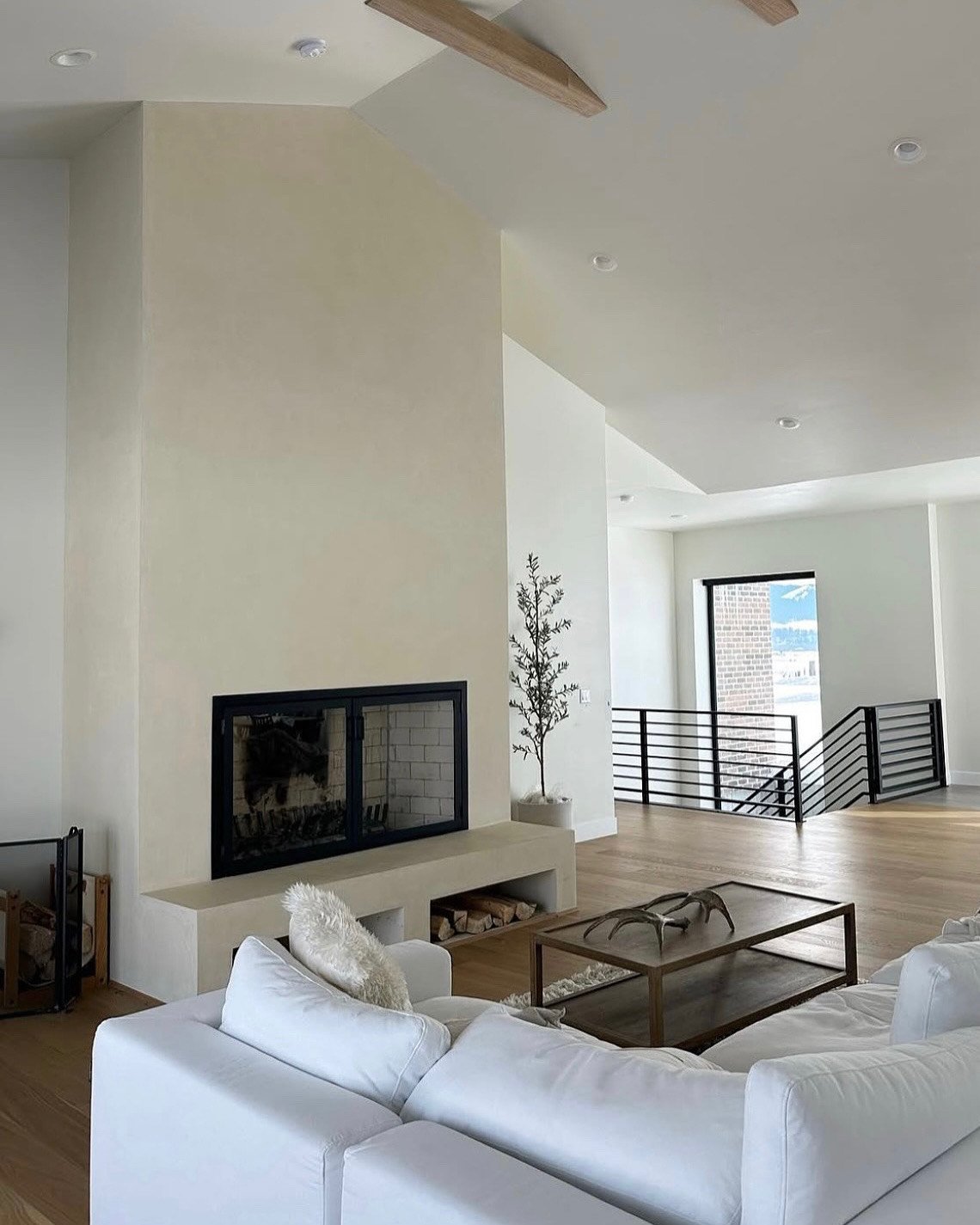 Modern living room with white walls and ceilings, featuring a minimalist fireplace, white sofa, glass coffee table, and wooden flooring. Large windows and railing leading to stairs in background.