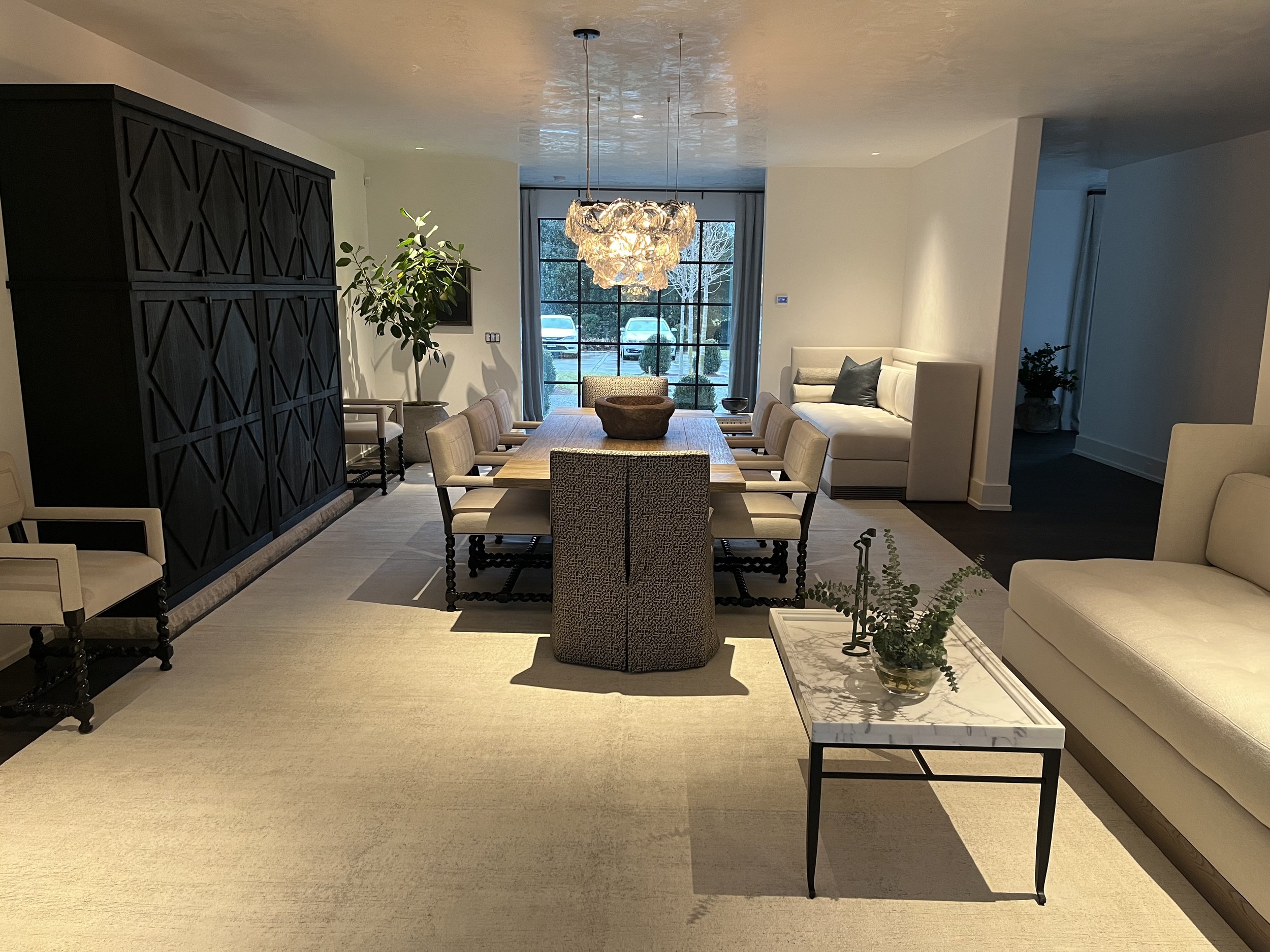 Modern living and dining room with beige and black decor, featuring a large wooden dining table with chairs, a white sofa, a dark wood cabinet, pendant lighting, and green plants.