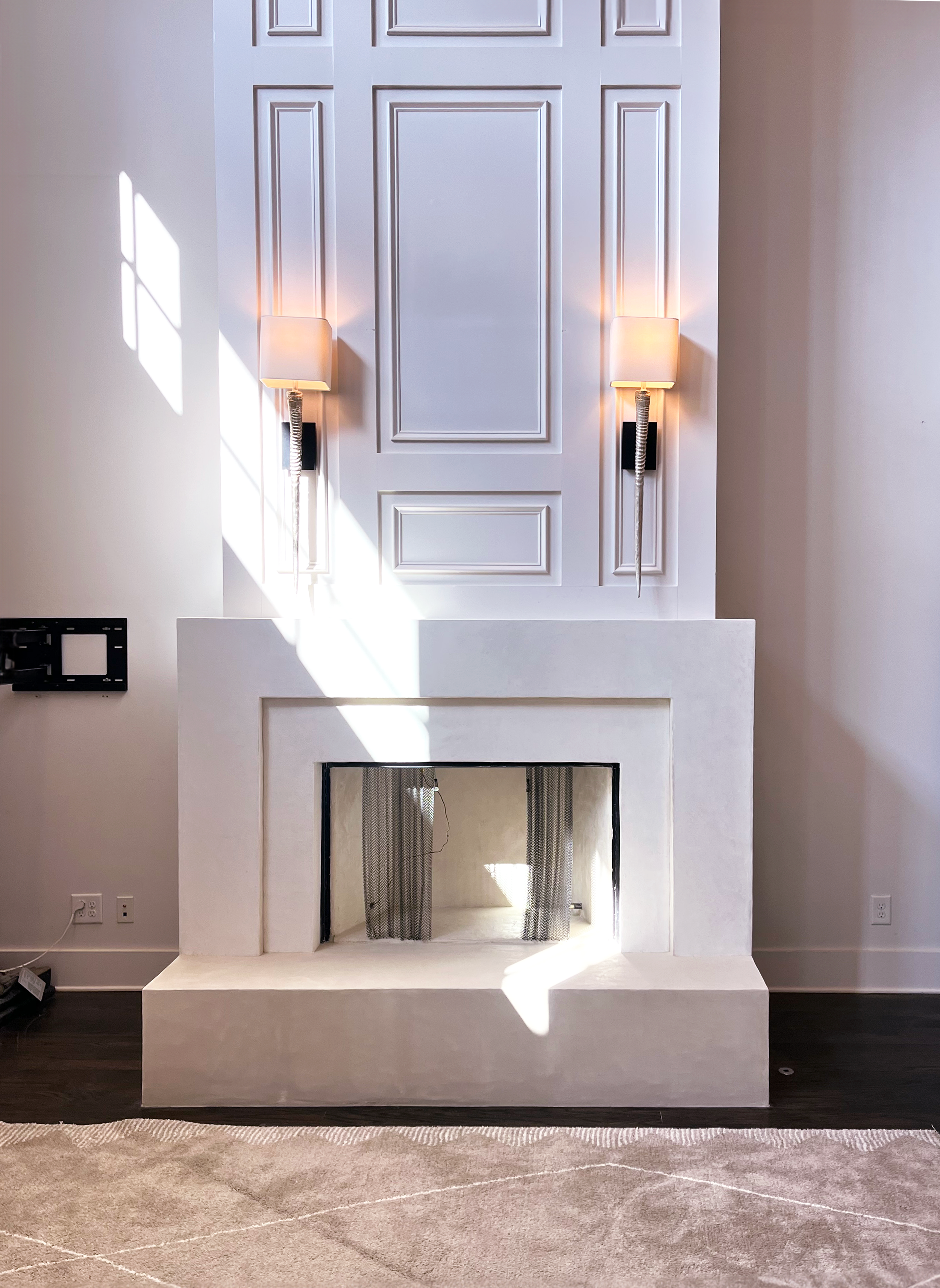 White modern fireplace with panel detailing, two wall sconces, sunlight casting shadows, and a carpeted floor.