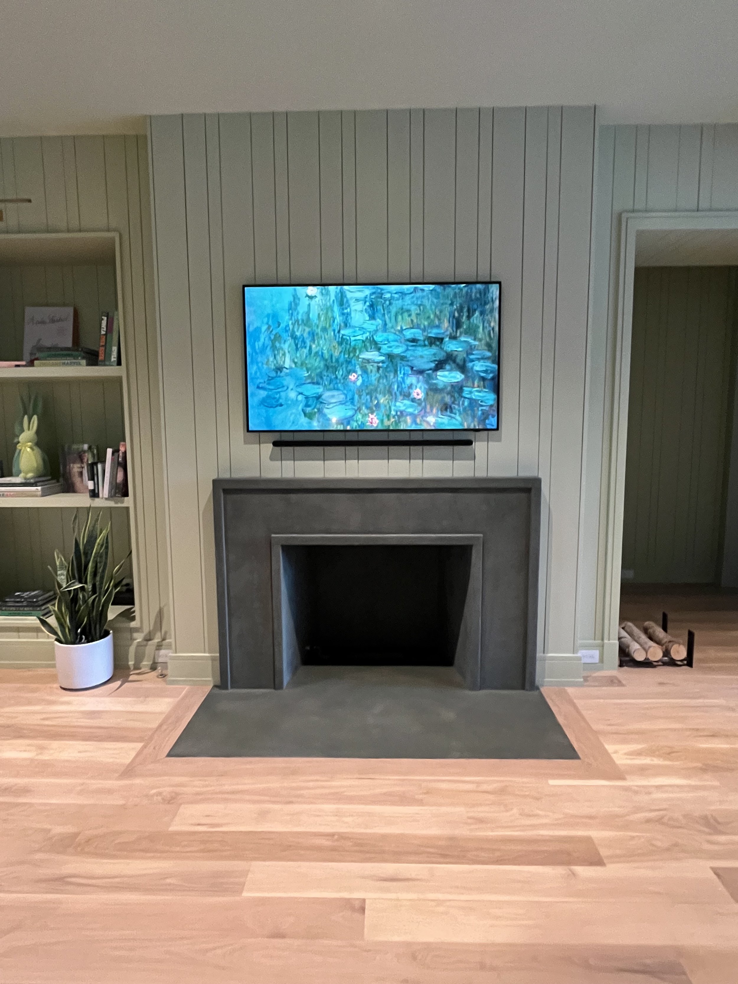 Modern living room with minimalist fireplace, flat-screen TV displaying a water lily painting, wooden floor, potted plant, and bookshelf on the left. Firewood is neatly stacked on the right.