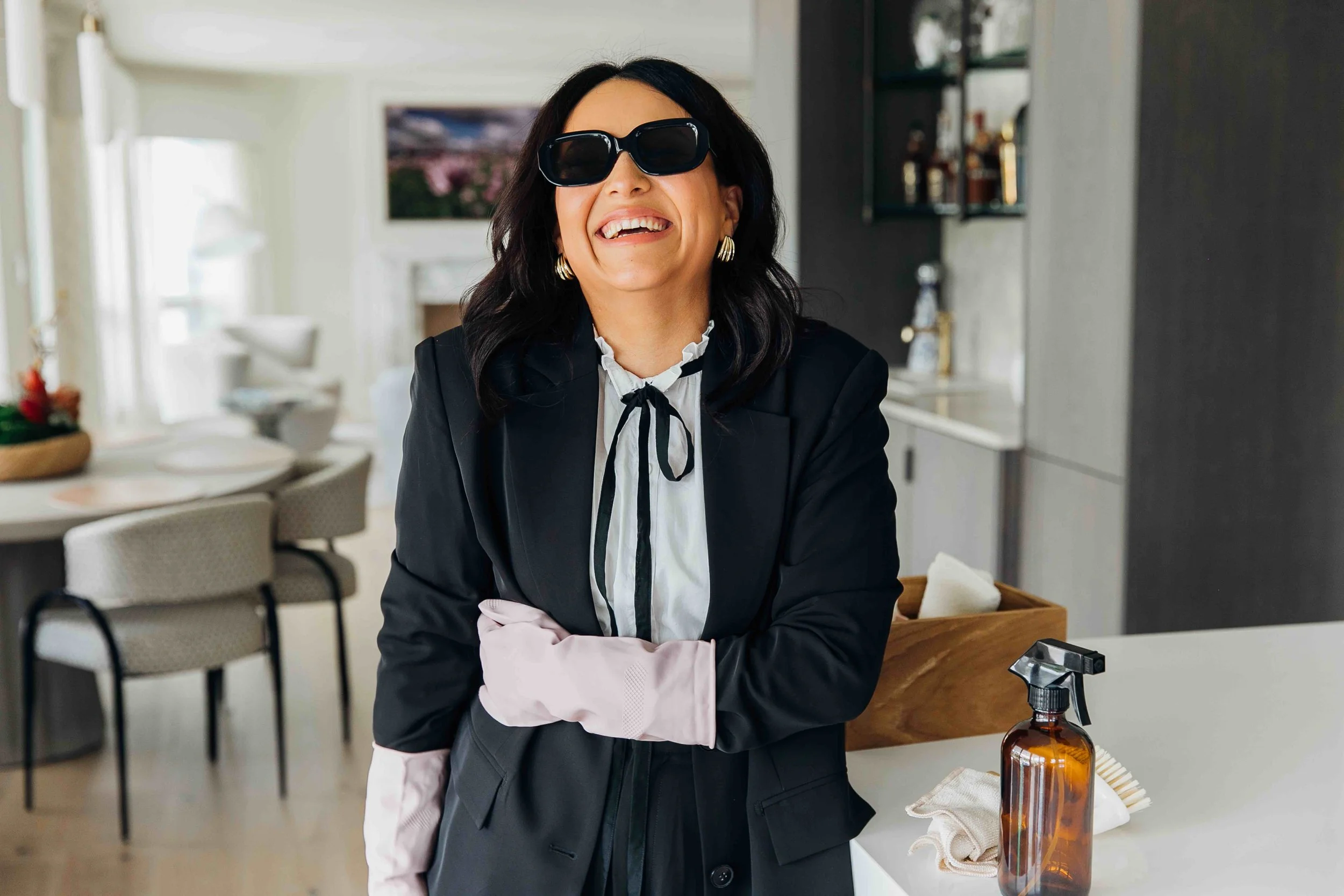 A woman with dark hair wearing sunglasses, a black blazer, and pink gloves smiling in a modern kitchen.