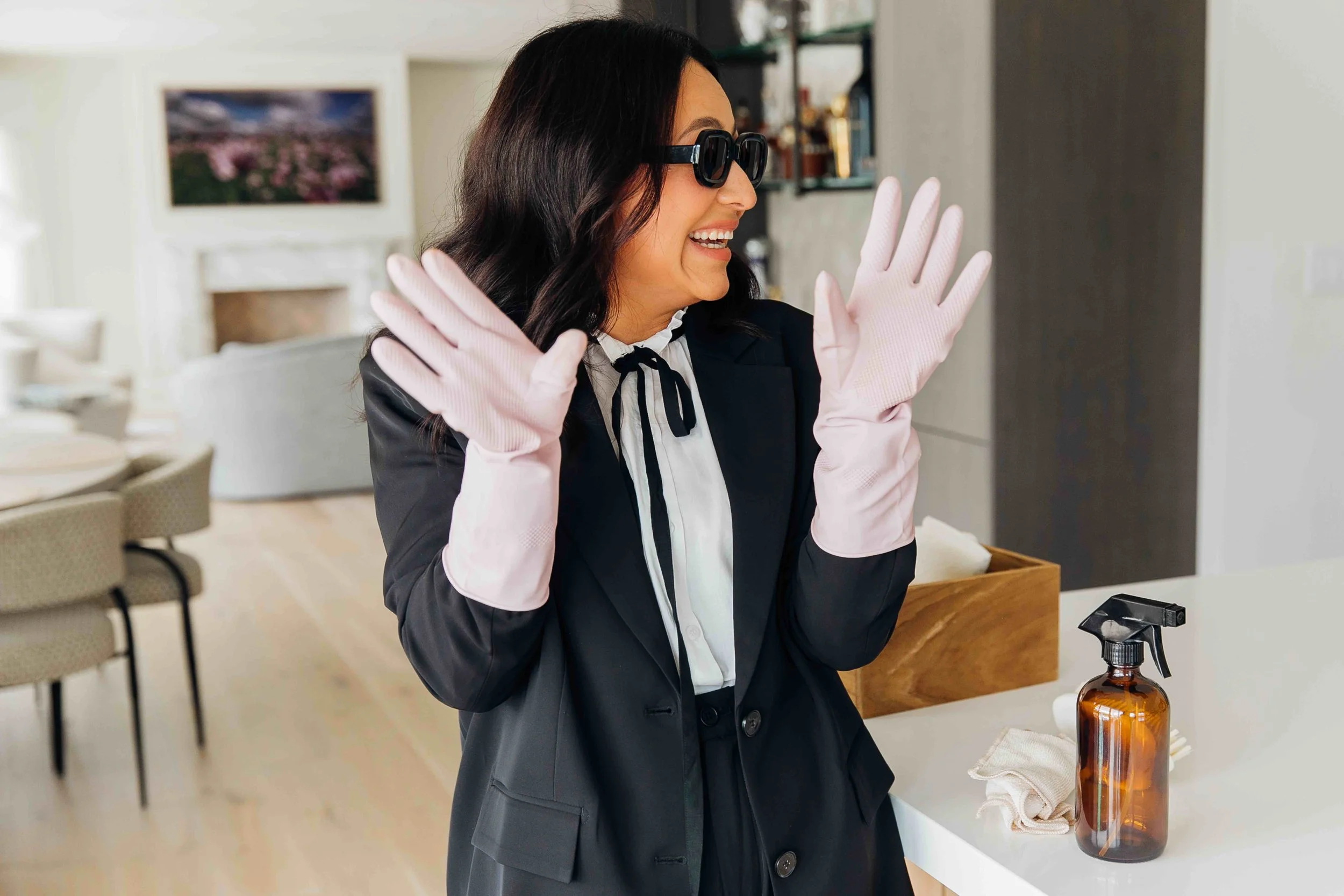 Woman in black blazer and pink cleaning gloves smiling and gesturing with hands, standing near a cleaning spray bottle and cloth on a white countertop in a modern living room.