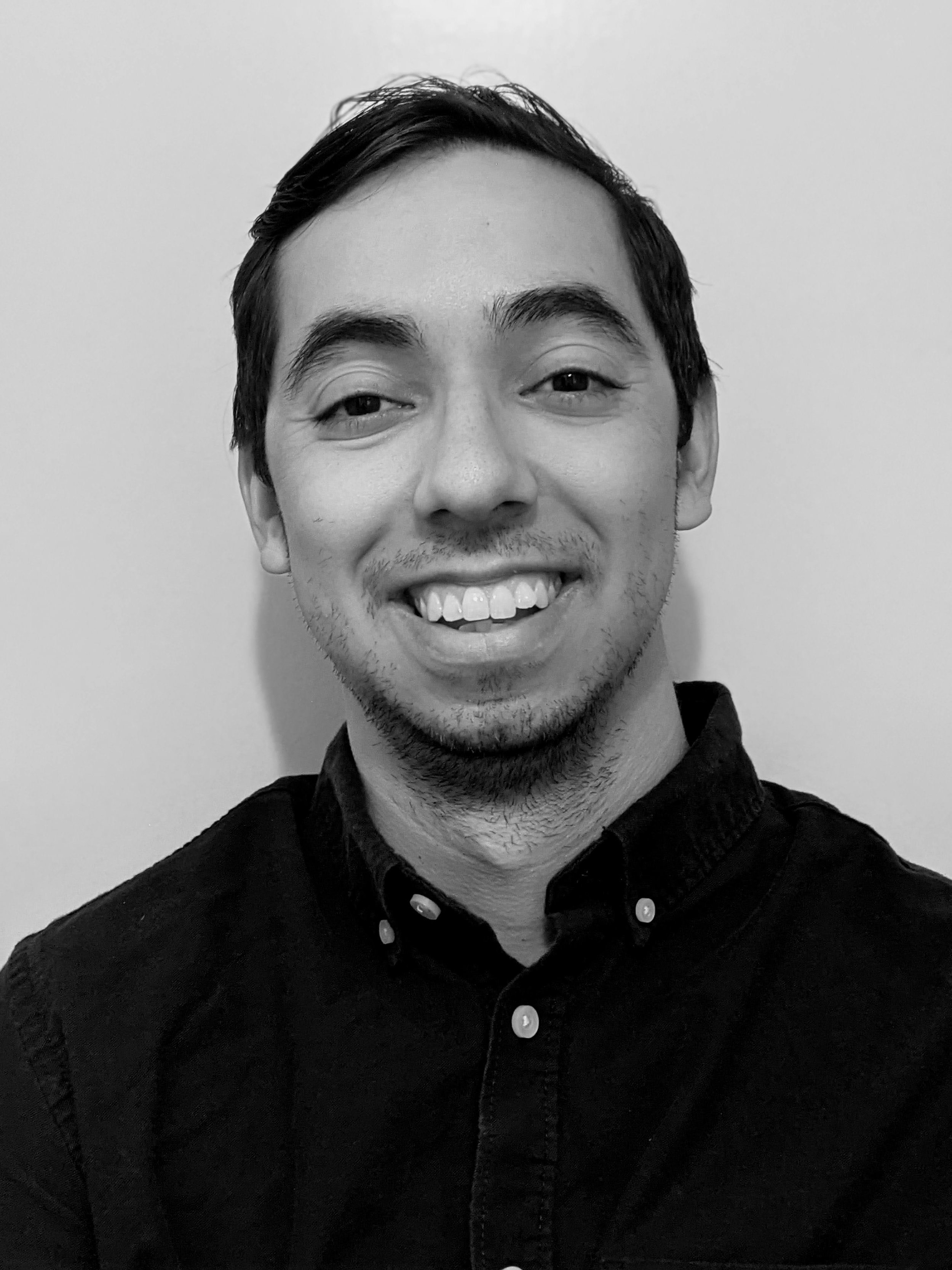 Black and white portrait of a smiling young man with dark wavy hair, wearing a zip-up jacket over a collared shirt, standing next to a window with multiple small panes.