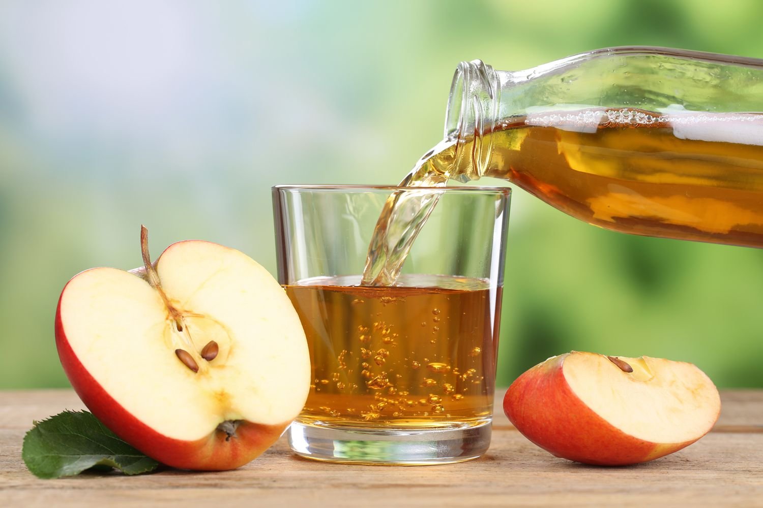 A glass of apple juice being poured from a glass jar, with a sliced apple on a wooden surface, and a blurry green outdoor background.