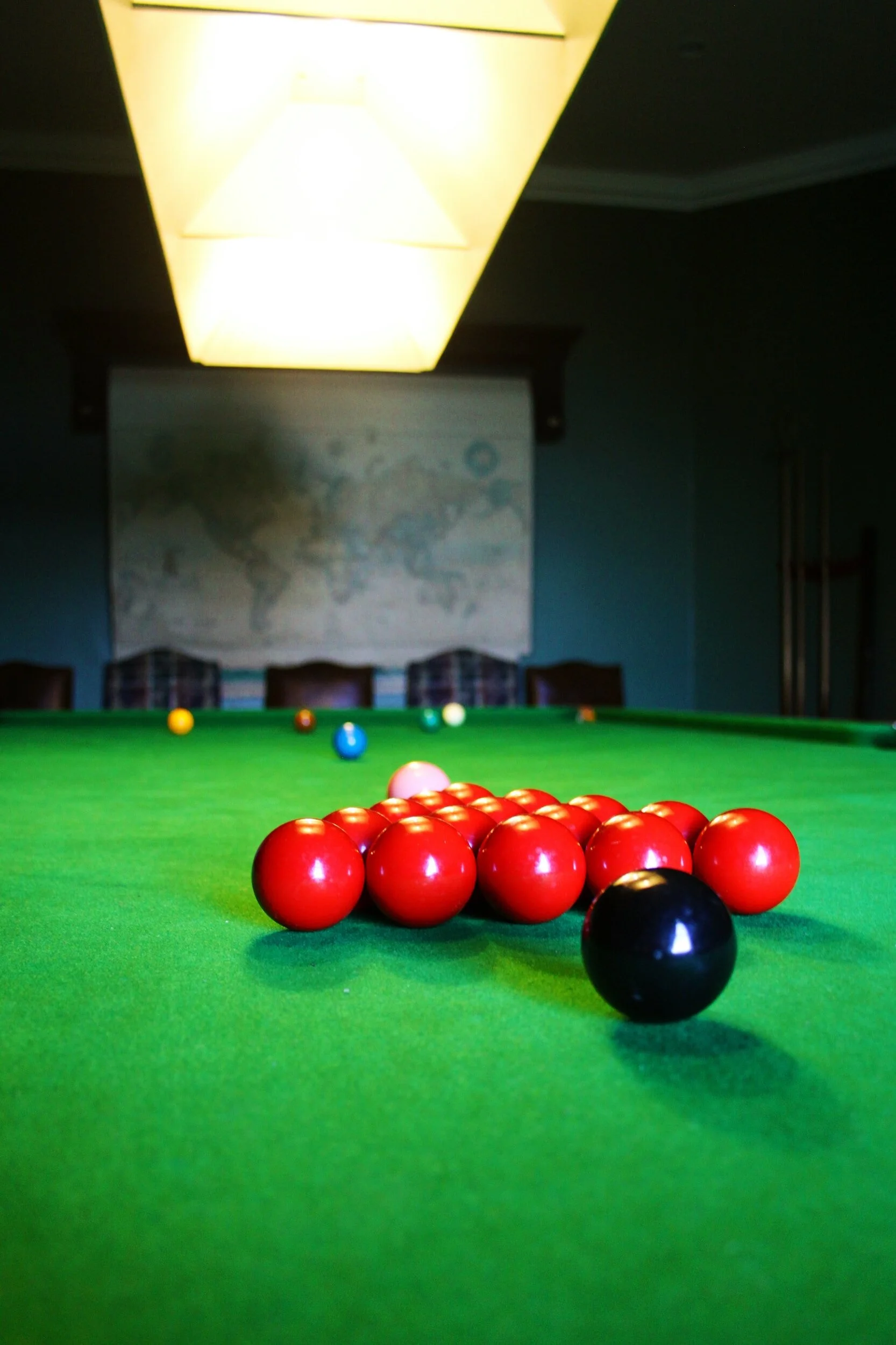 A billiard table with red balls and a black ball in a dimly lit room, with a ceiling light and a world map on the wall in the background.