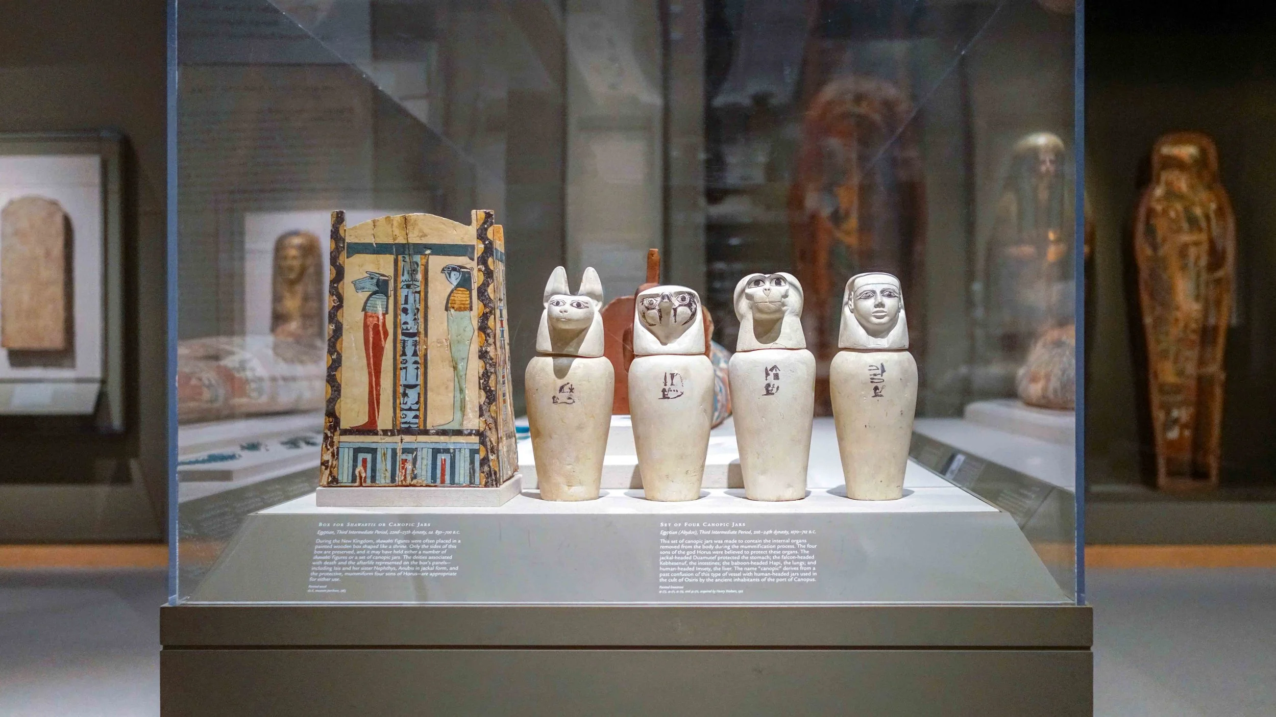 Ancient Egyptian canopic jars and painted wooden box displayed in a museum case. The jars are shaped like human and animal heads, with hieroglyphs etched on them, and are positioned in front of a painted wooden box with Egyptian motifs.