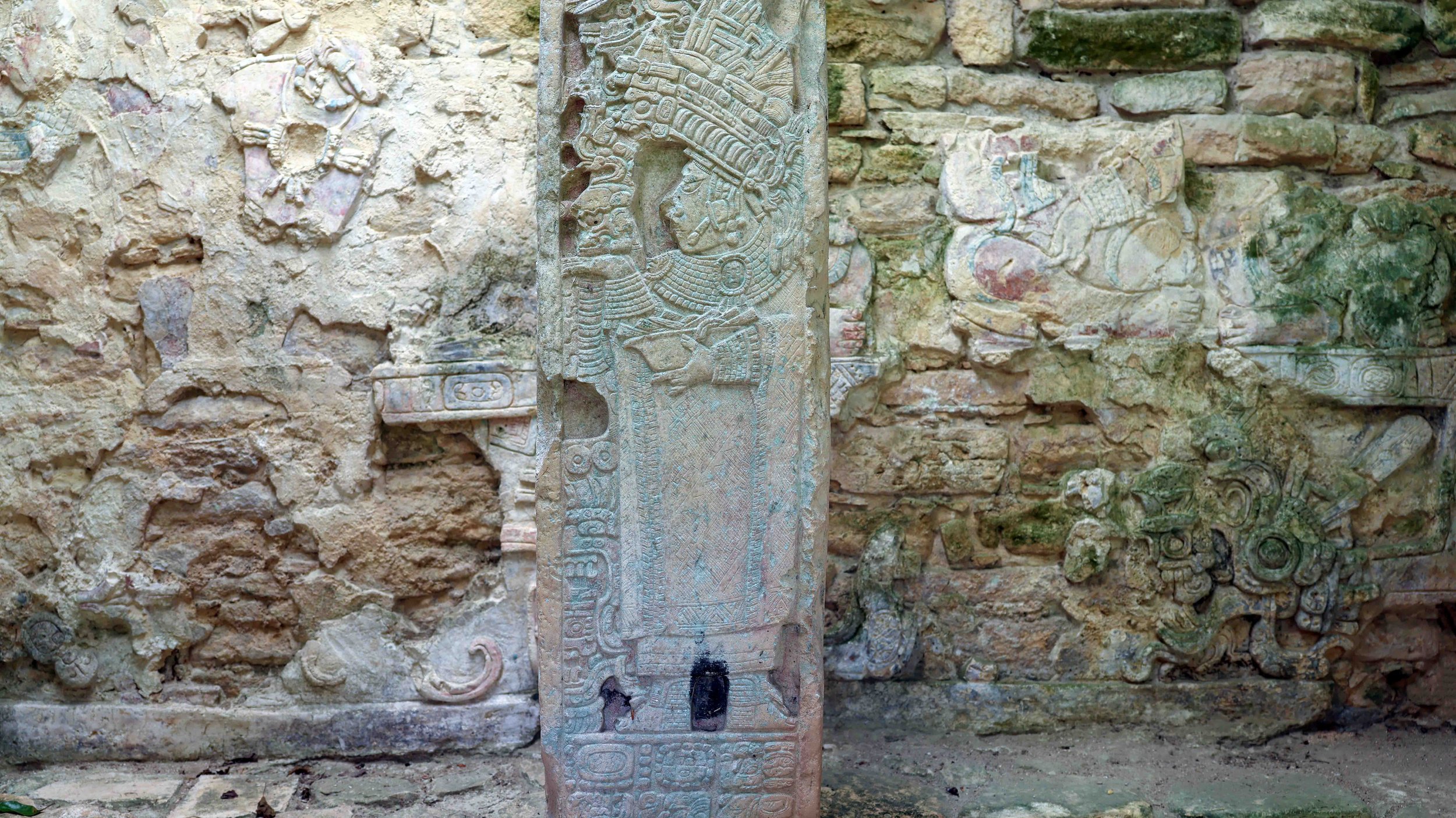 Ancient stone carvings of Mayan figures and symbols on a wall and a central stone slab in a historical archaeological site.