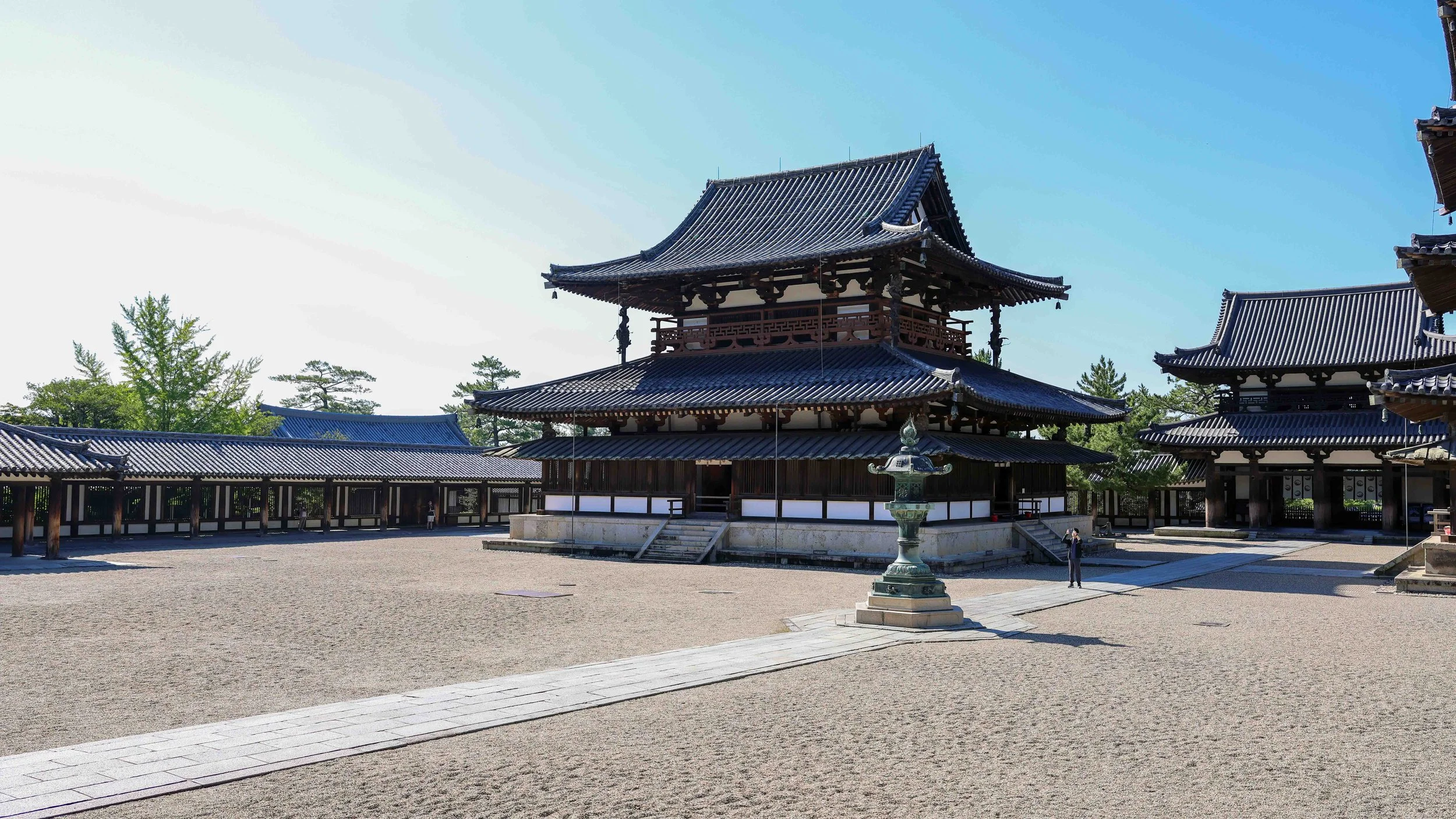 Traditional Japanese temple with wooden structures and tiled roofs, surrounded by trees and a stone courtyard with a lantern in the foreground.