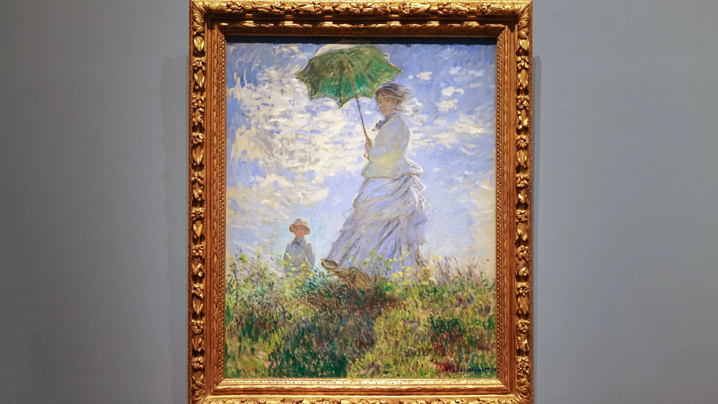 Impressionist painting of a woman holding a green parasol and a young girl walking through a field of yellow flowers under a partly cloudy sky, framed in an ornate gold frame.