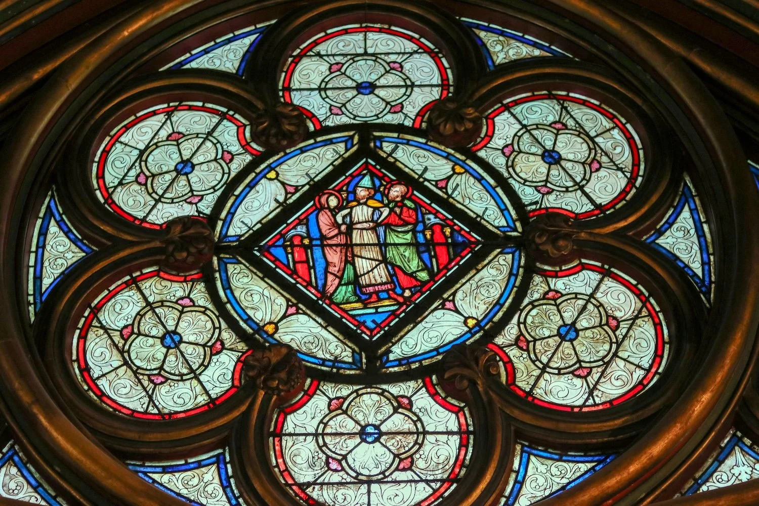 Intricate stained glass window depicting three figures in a geometric design, surrounded by floral patterns and colorful detailing in blue, red, and green hues.