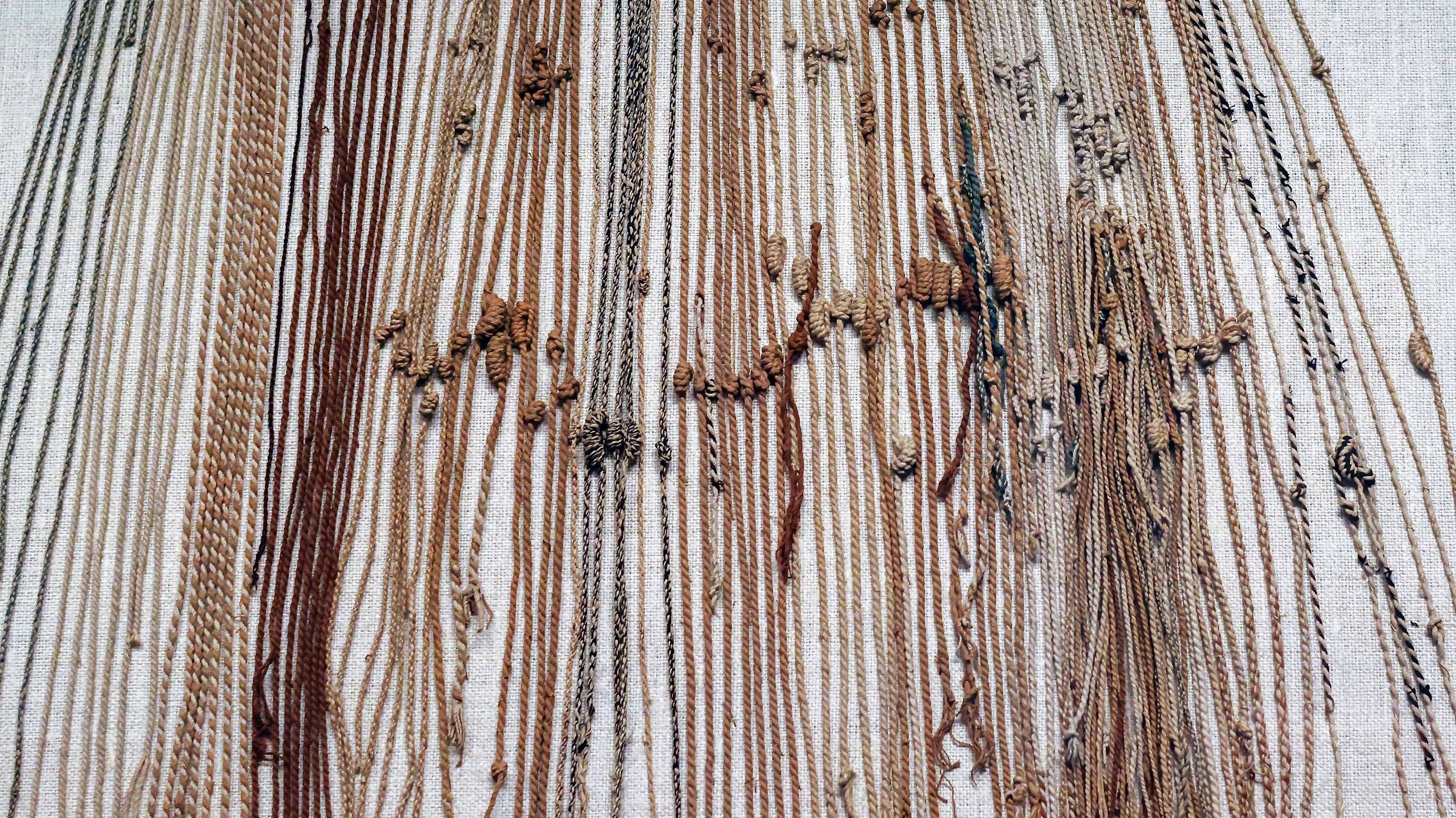 A woven textile with various brown, beige, and black threads, with knots across it.