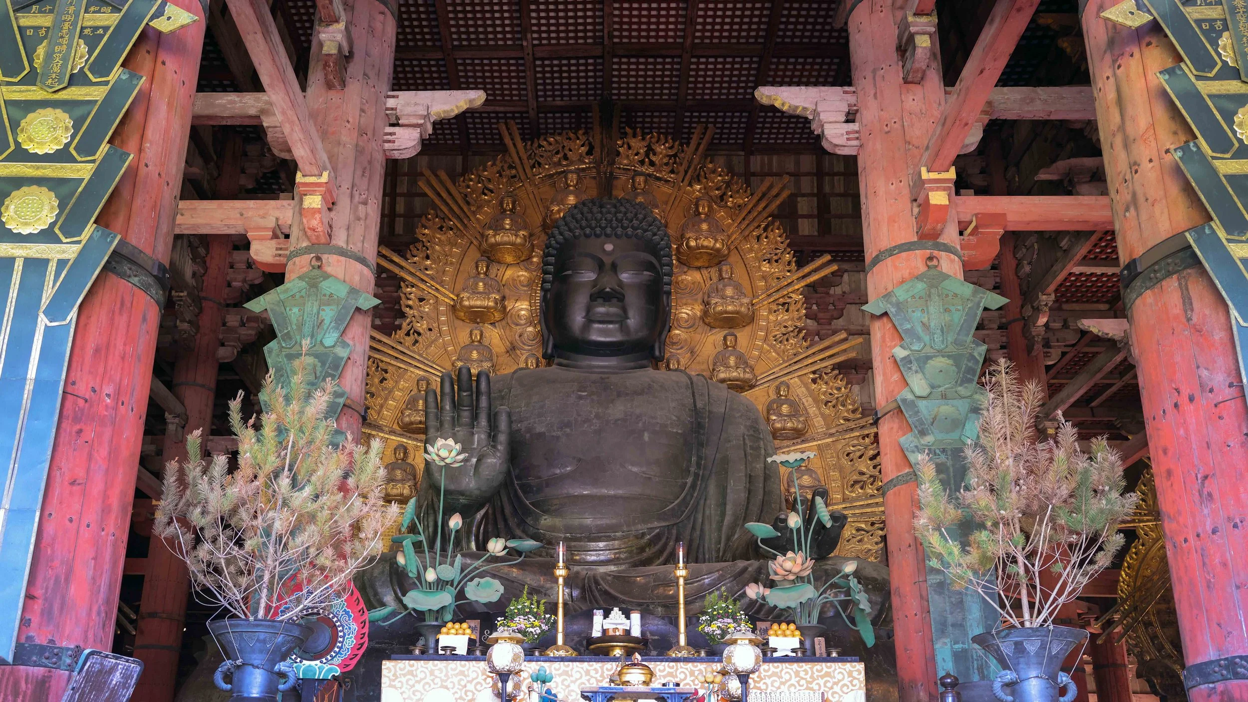 A large black Buddha statue seated indoors, with a gilded halo and smaller statues behind, surrounded by flowers, candles, and traditional decorations.