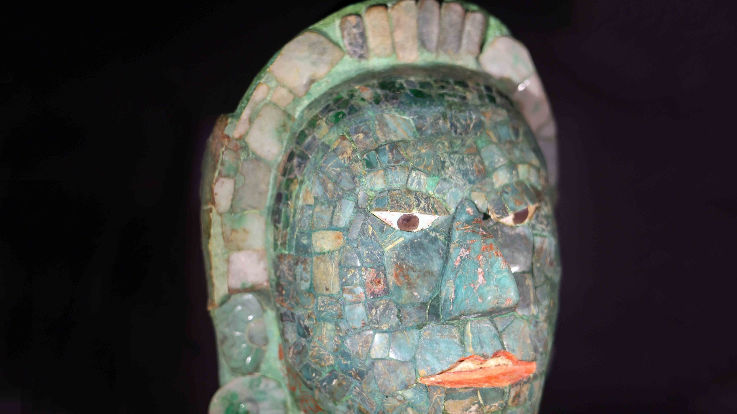 Ancient mosaic face sculpture with green, blue, and red tiles, set against a black background.