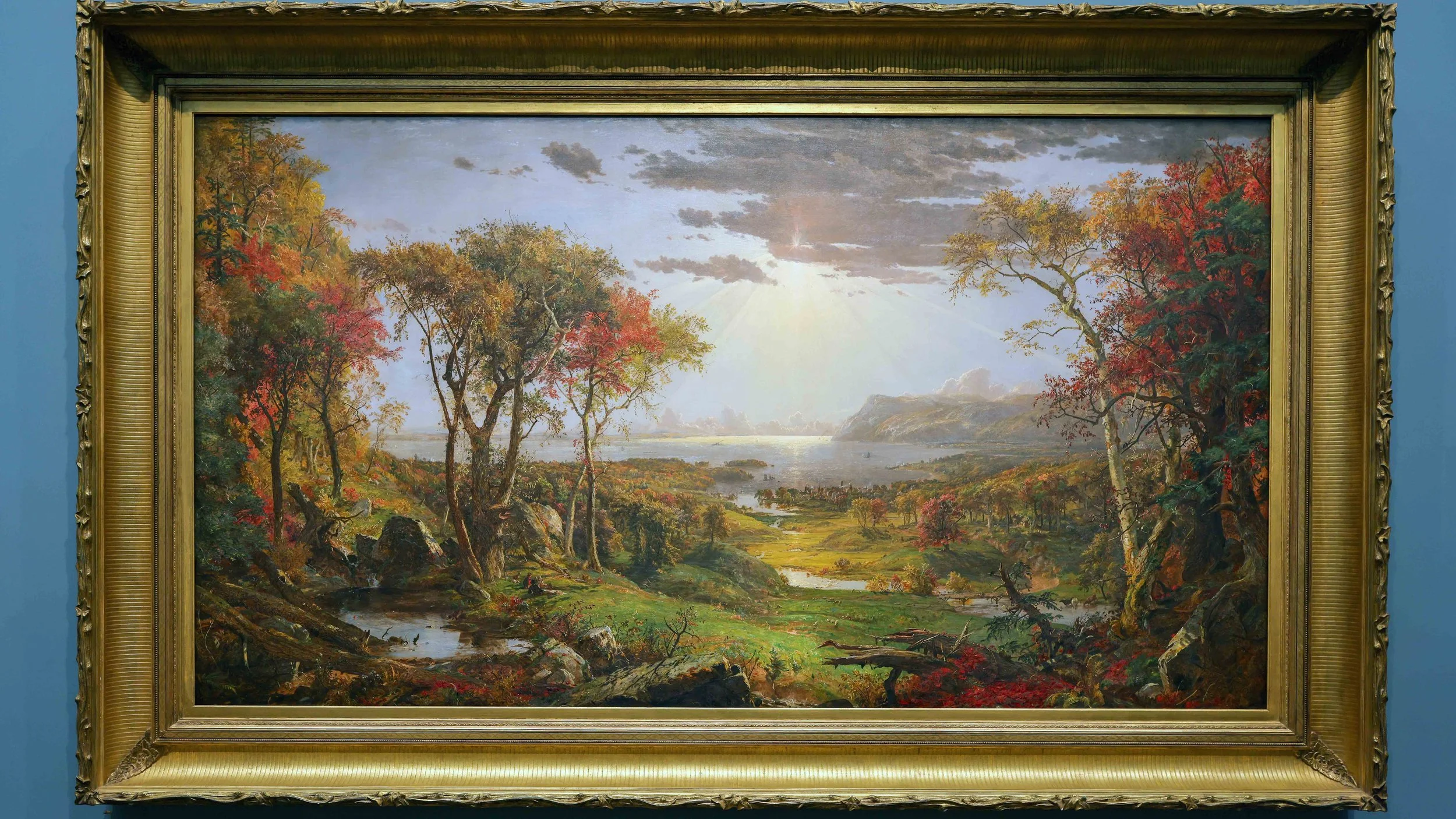 Landscape painting of trees with autumn colors, a stream, and a distant river or lake under a sky with clouds and sunlight.