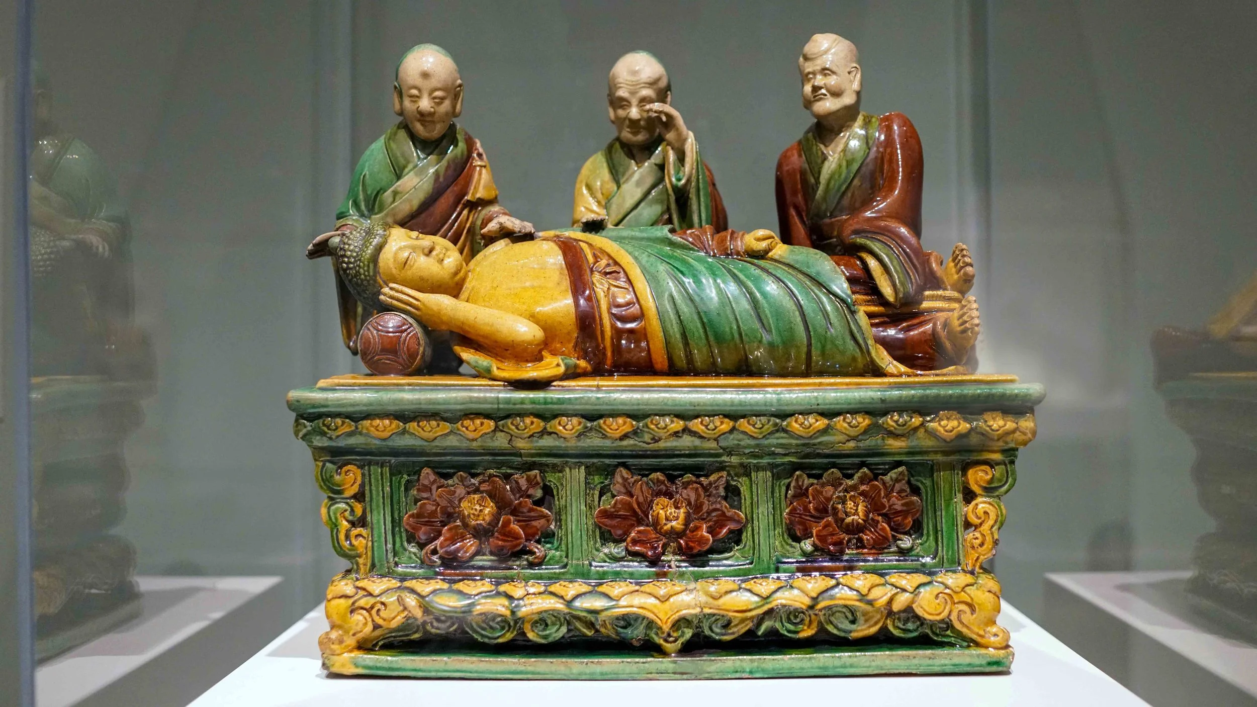 Colorful ceramic sculpture of three monks surrounding a reclining figure, displayed in a glass case.