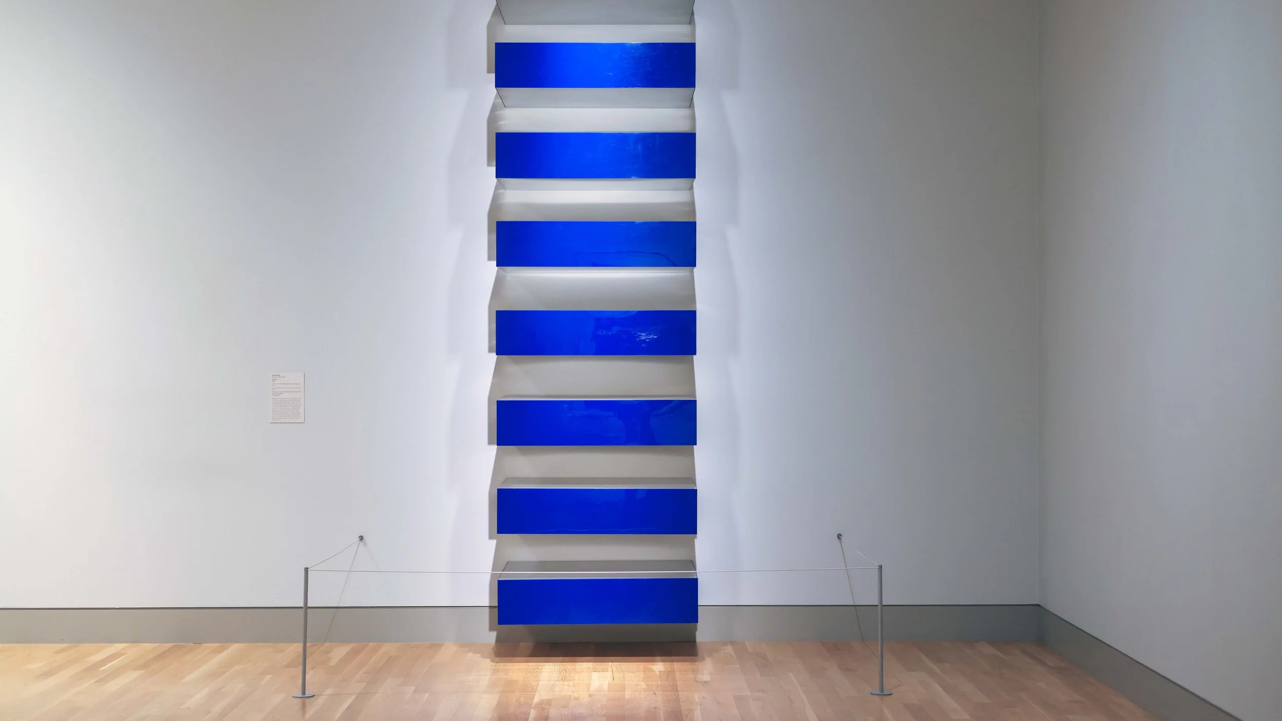 A modern art installation with alternating horizontal blue and white panels, arranged vertically on a white wall in a gallery.