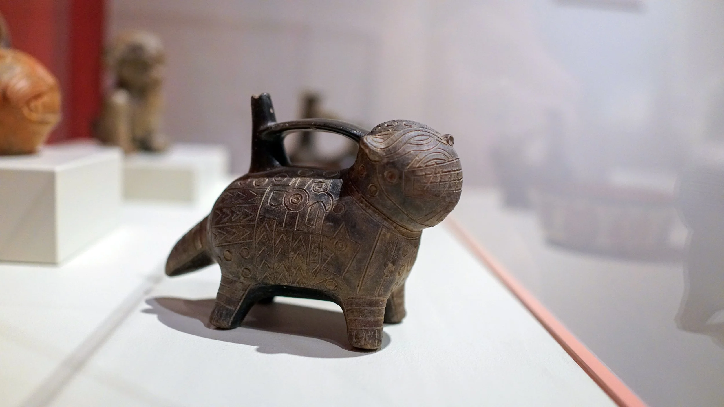 A carved figurine of an animal, possibly a boar or pig, with intricate geometric patterns on its body, displayed in a museum or gallery setting.