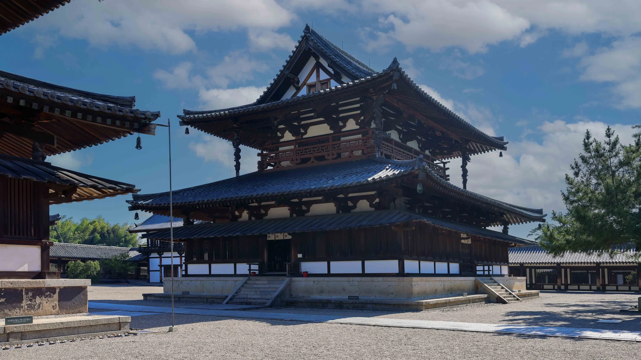Traditional Japanese wooden temple with multiple tiered roofs, set outdoors under a partly cloudy sky, surrounded by trees