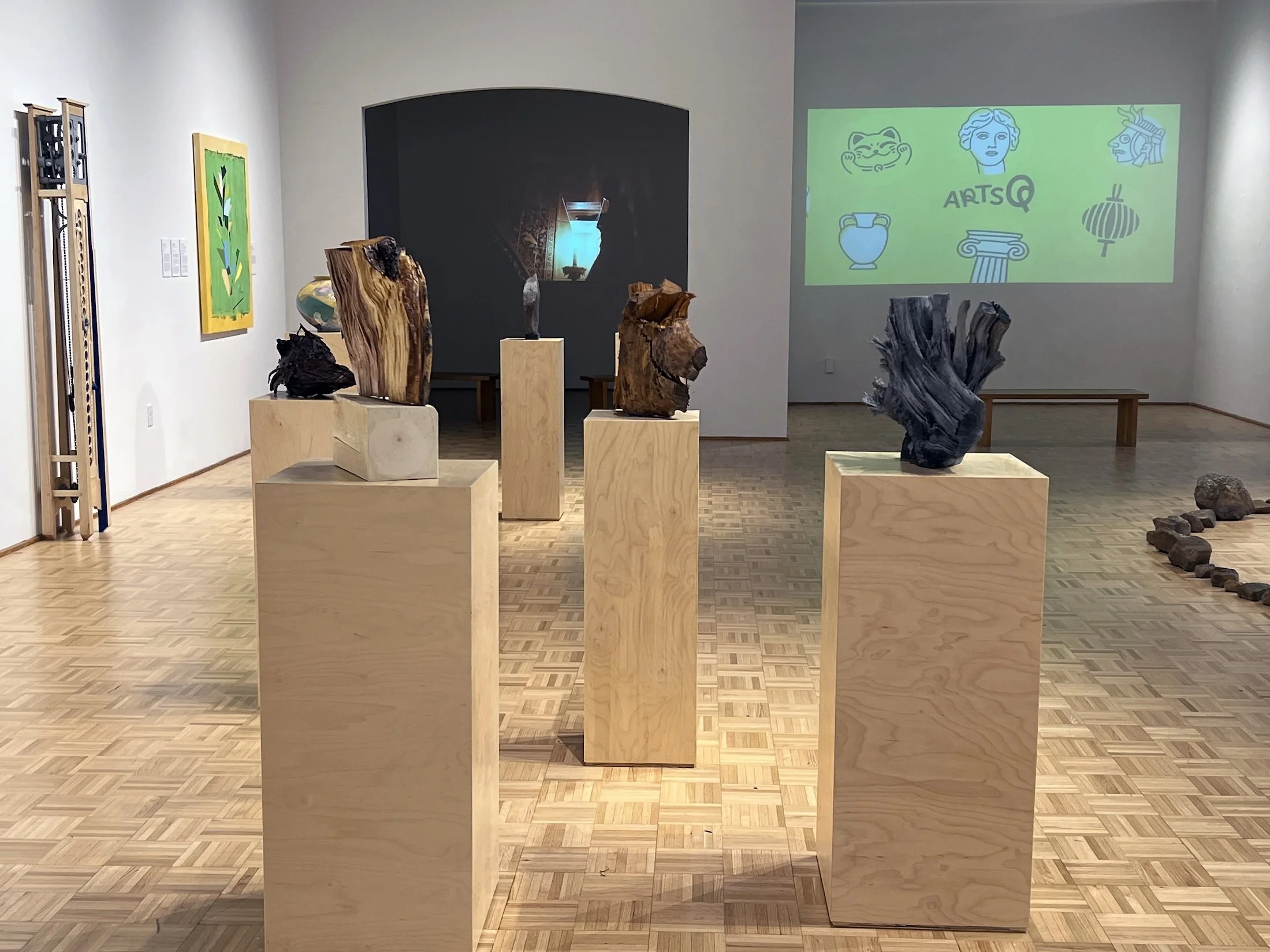 Art gallery with wooden sculptures displayed on light wood pedestals, a colorful painting on the wall, and a digital screen showing art-related icons and text.