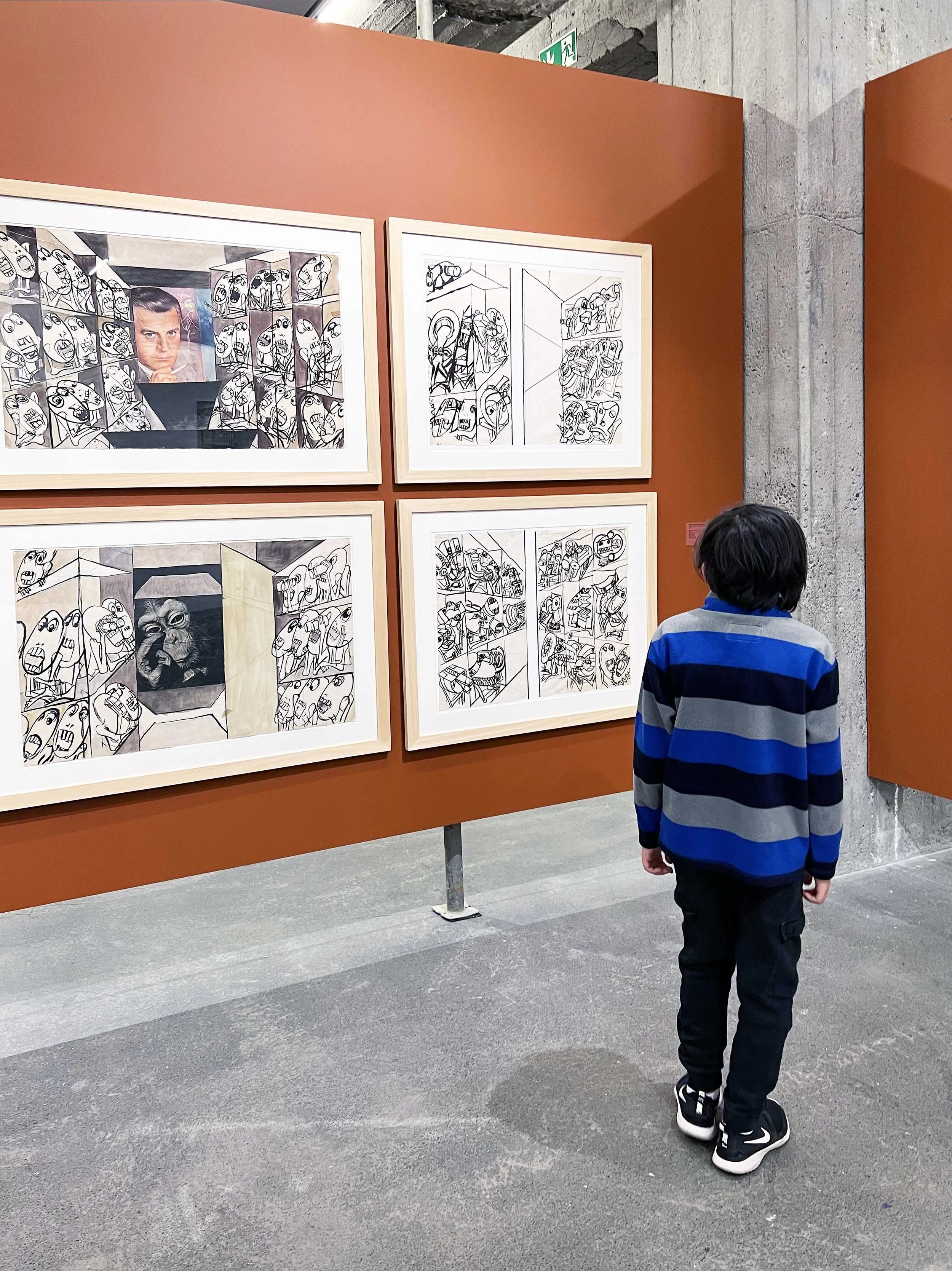 A young boy in a blue and gray striped sweater stands in front of an art gallery wall with framed black and white and colored artworks, observing the pieces.