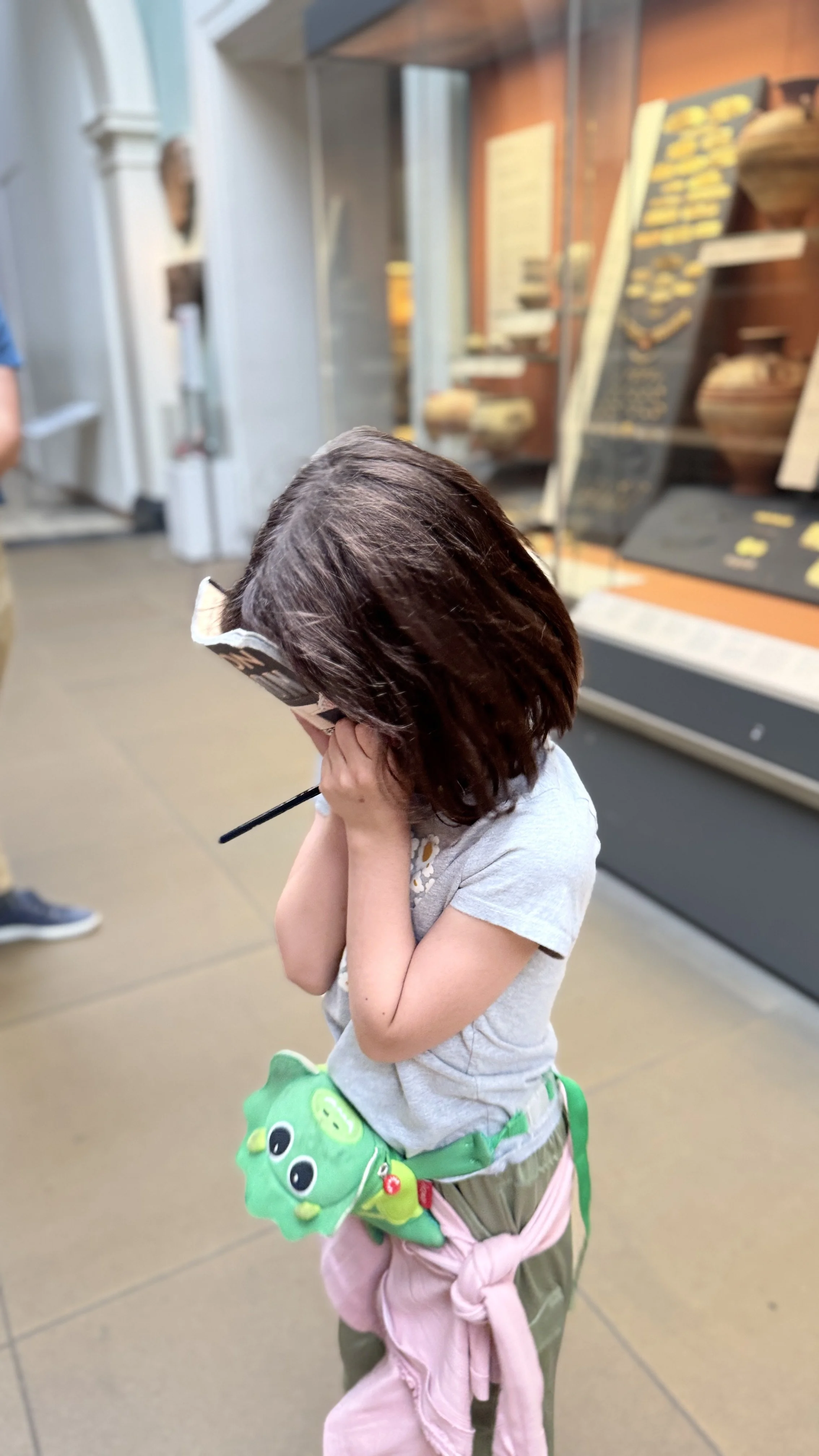 A young girl with brown hair covering her face, standing indoors near a display case, holding her head with both hands. She is wearing a gray t-shirt, a green purse with a cartoonish face, and pink jacket tied around her waist.