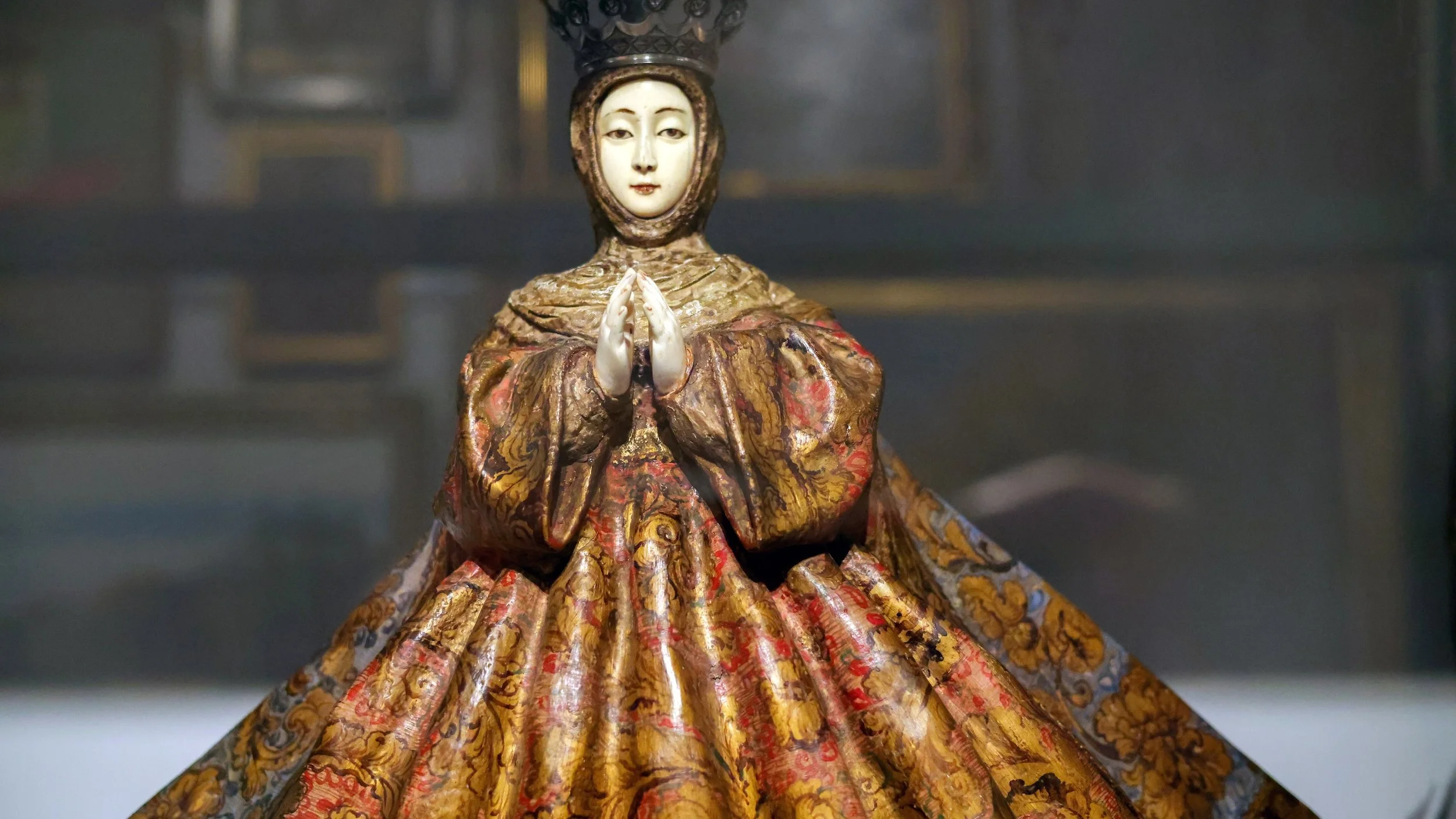 A religious statue of a woman in ornate clothing with hands in prayer, standing in a dimly lit room.