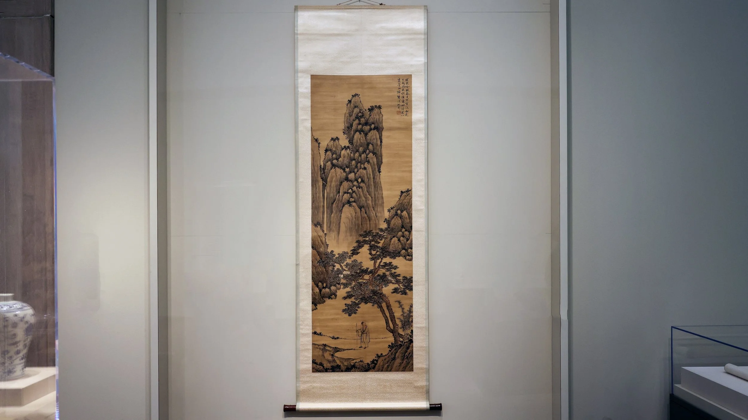 A traditional Chinese landscape hanging scroll with mountains, trees, and a person, displayed on a white wall in a gallery or museum.