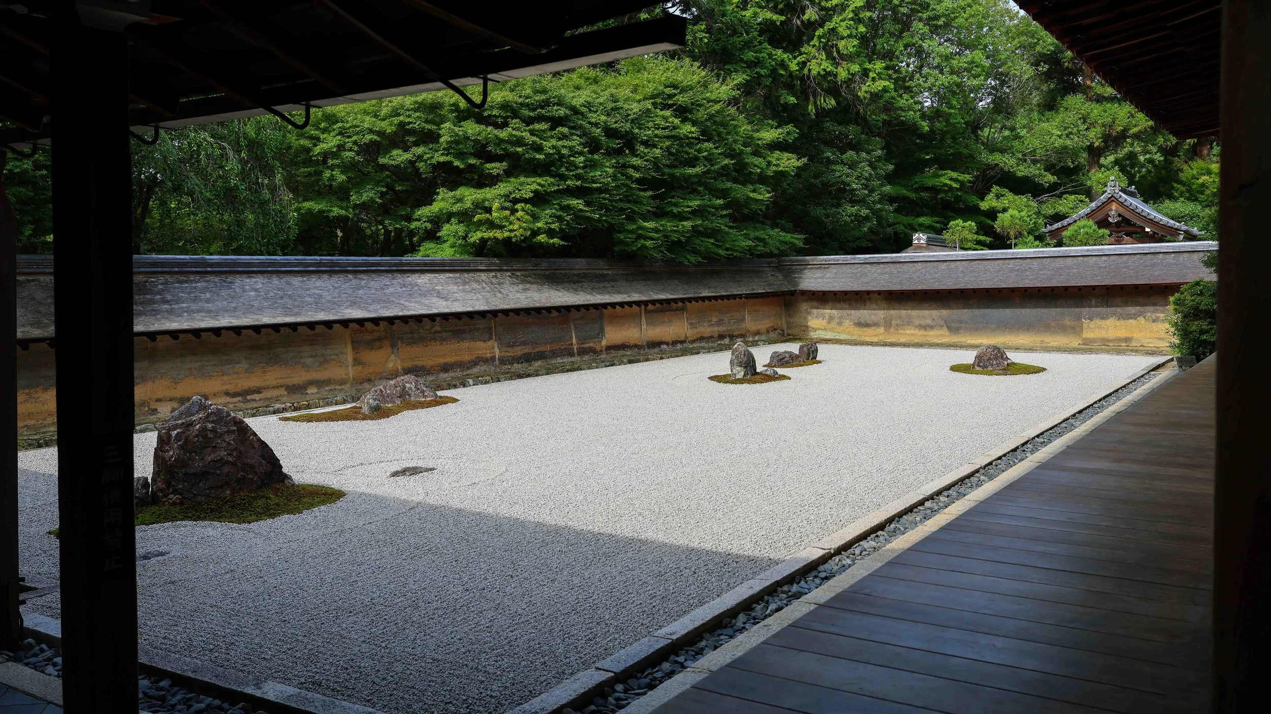 A traditional Japanese rock garden with raked white gravel, large rocks arranged on moss patches, surrounded by a wooden fence and lush green trees, viewed from a shaded veranda.