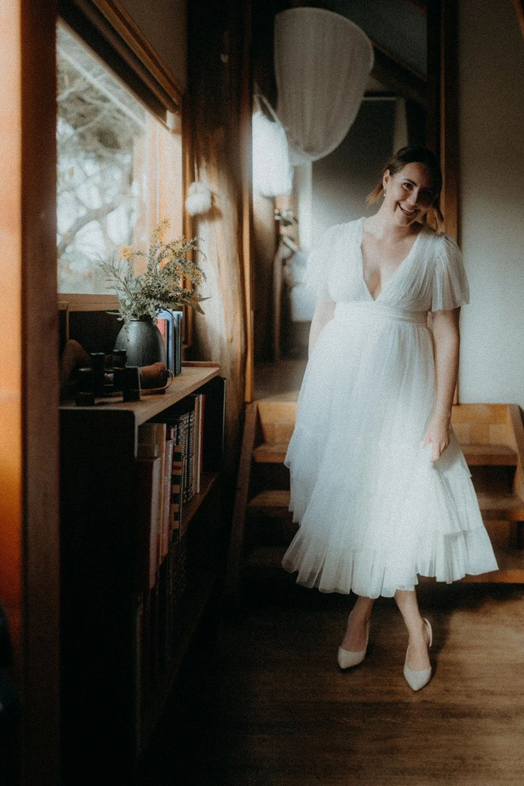 A woman in a white dress smiling and standing inside a room near a window with wooden trim and a bookshelf with books and a vase with flowers.
