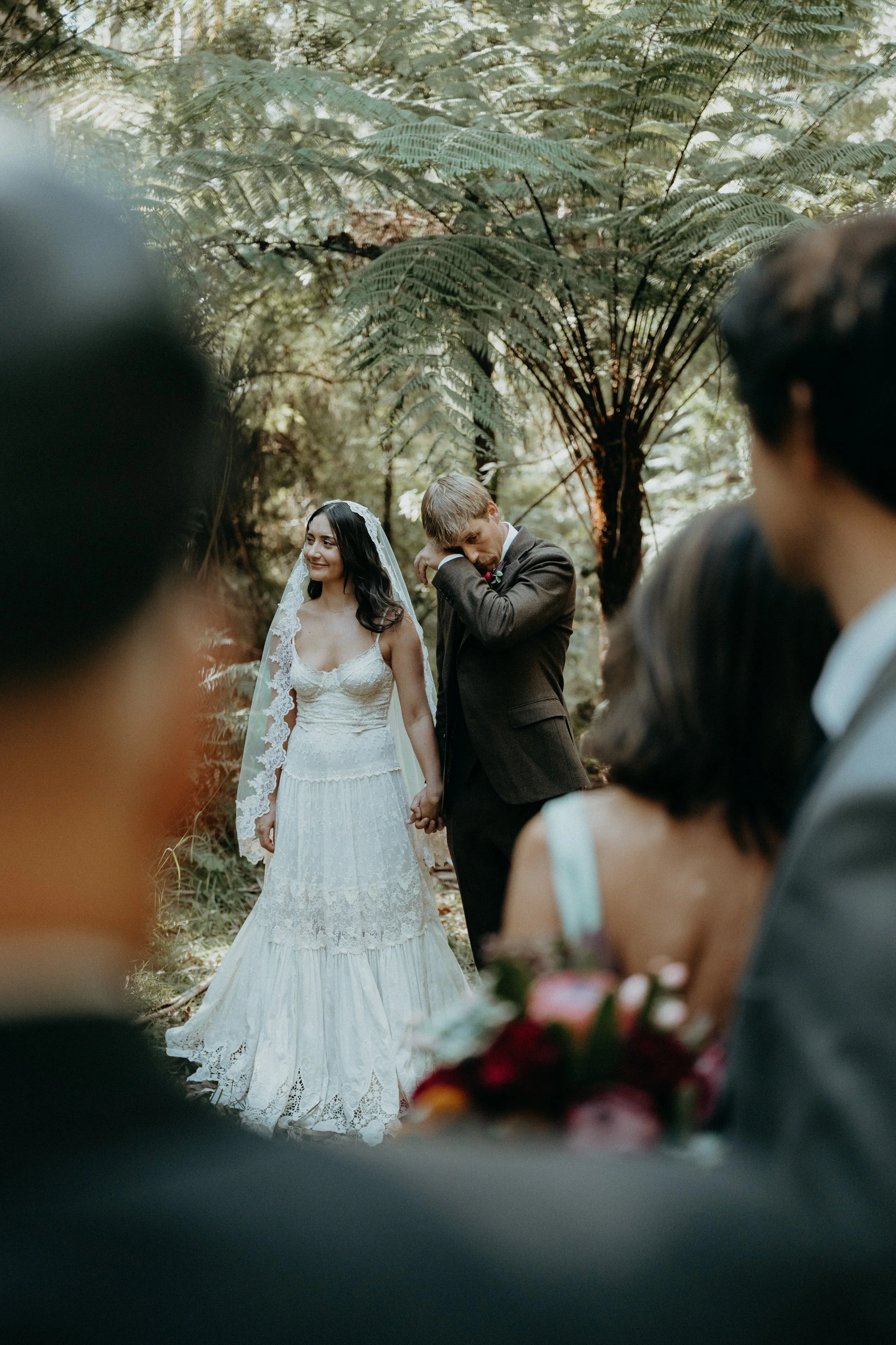A bride and groom holding hands during an outdoor wedding ceremony surrounded by greenery, with guests watching in the foreground.
