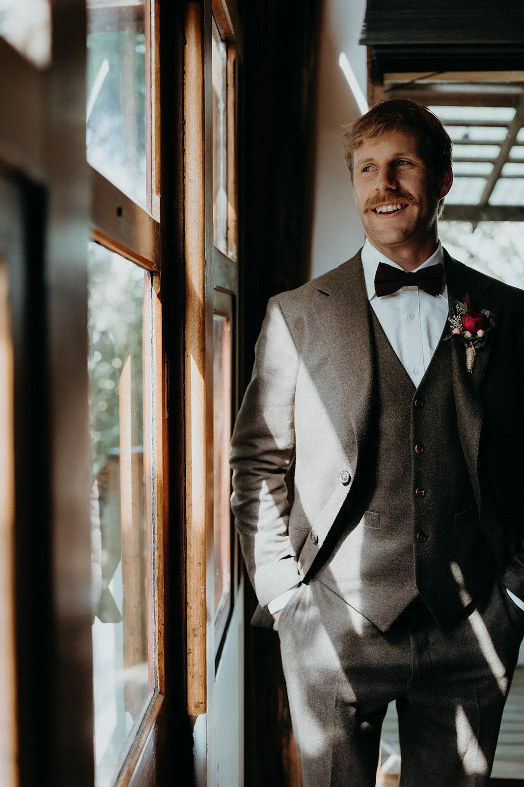 A man in a grey suit with a white shirt, black bowtie, and boutonniere, smiling and looking out the window while standing indoors.