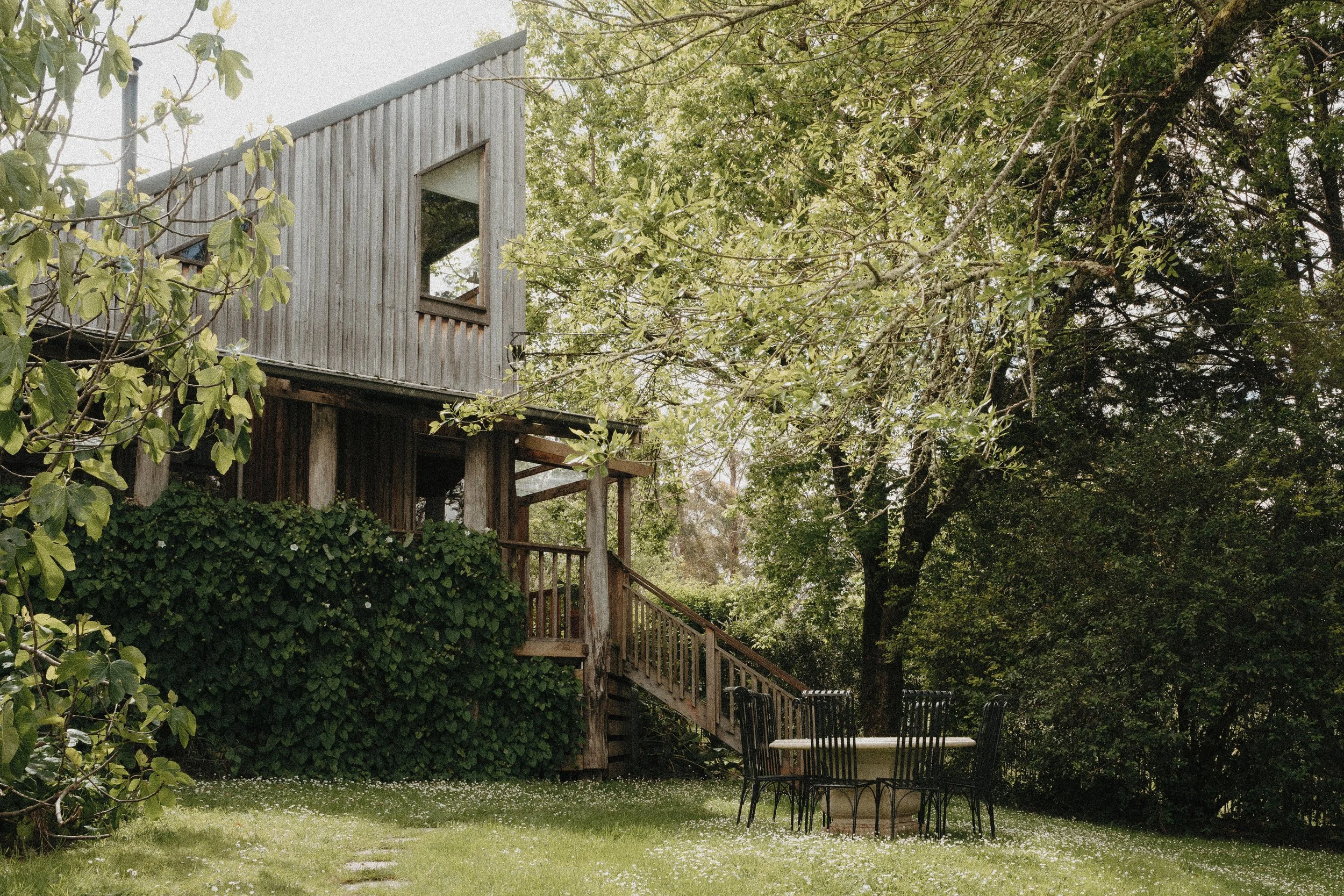 Outdoor backyard with a wooden treehouse, a black metal table, and chairs, surrounded by lush green trees and grass.