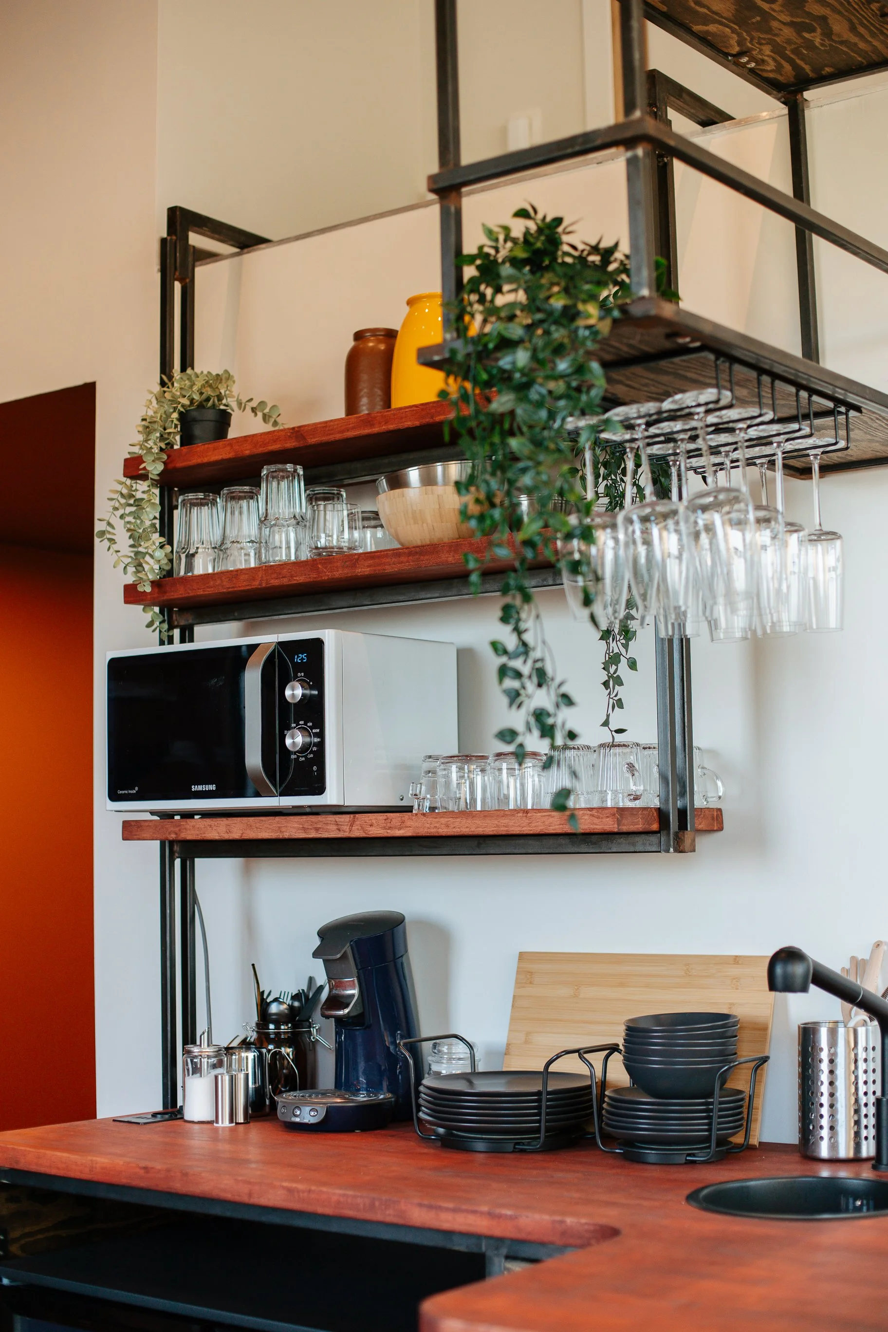Une cuisine moderne avec des étagères en métal et bois, un micro-ondes, une machine à café, des verres suspendus, et des vaisselle noire.