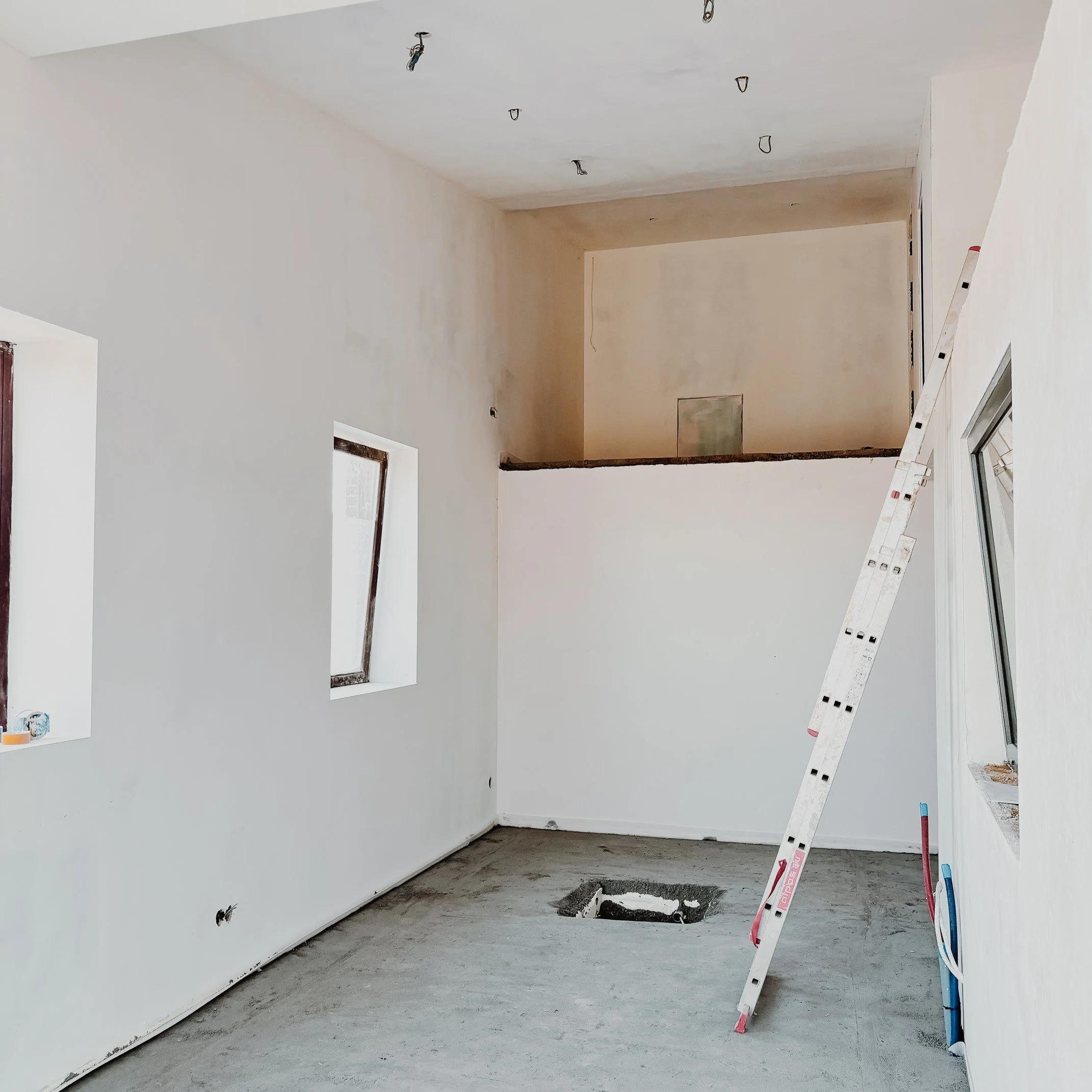 Pièce en construction avec murs et plafond blancs, fenêtre ouverte, escabeau en biais, trou au sol, câbles et éléments électriques.