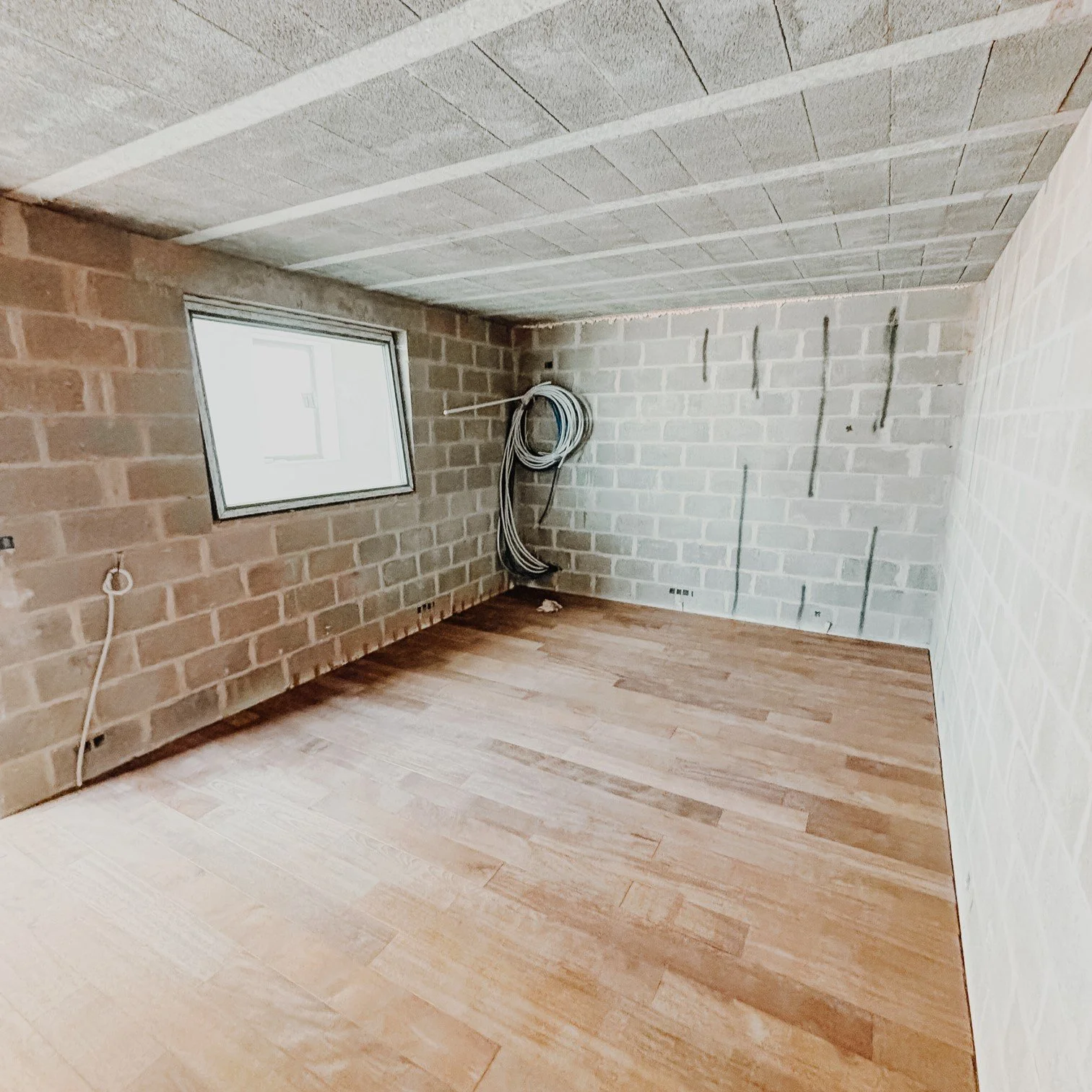 Une pièce vide en construction avec murs en briques et plafond en béton, une fenêtre sur le mur de gauche, des câbles électriques raccordés dans un coin, un sol en bois clair, et des marques de peinture ou de marquage sur le mur à l'arrière.
