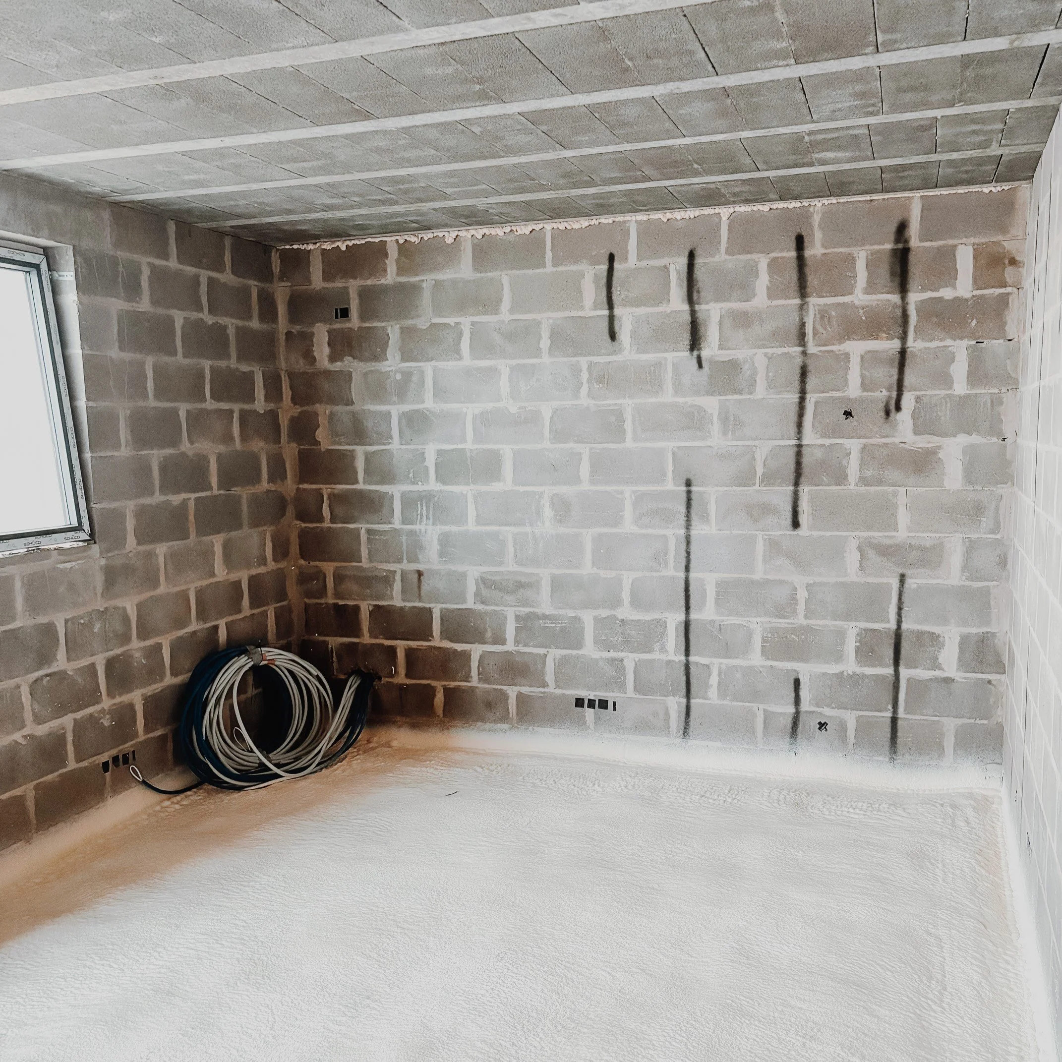 Une pièce en construction avec murs en brique apparente, un plafond en béton, un sol en béton fraîchement coulé, et des câbles électriques enroulés dans le coin inférieur gauche. Des marques de peinture noire en ligne verticale sont visibles sur le m