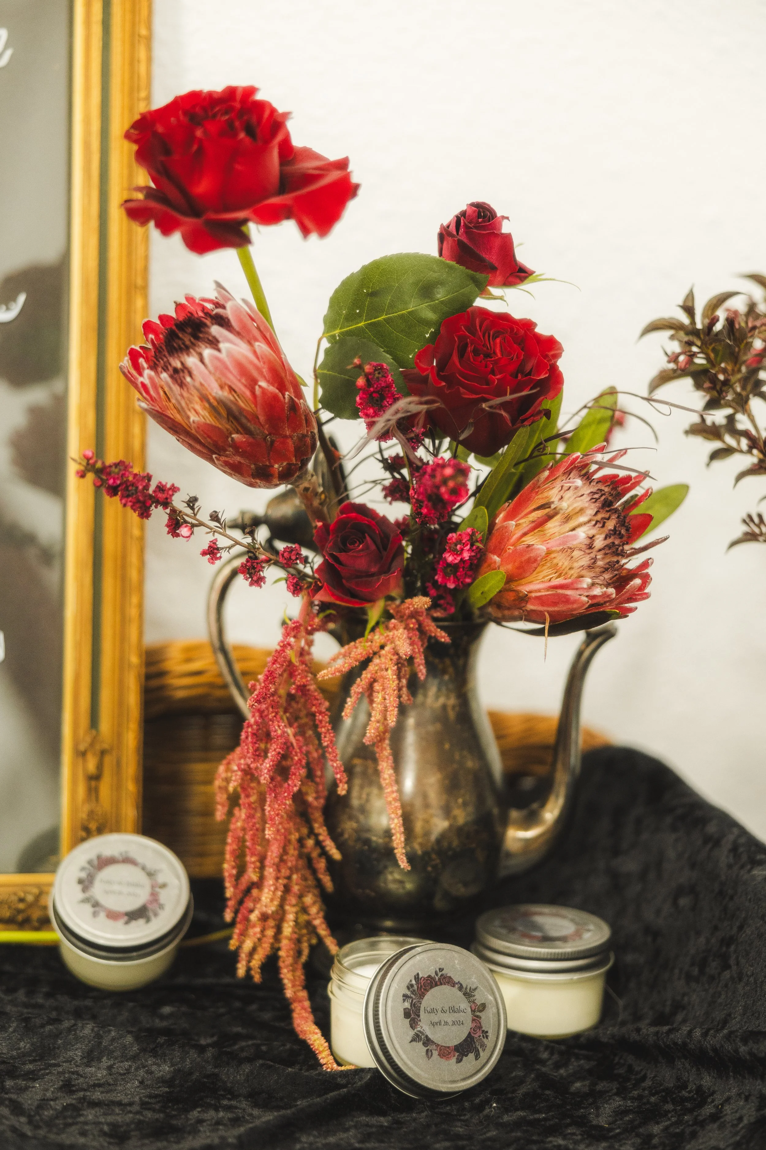 Floral "bud vase" using upcycled antique silver kettle. Custom labeled and scented candles for wedding guest gifts on wicker table and crushed black velvet drape from The Franklin Events and Community Center. 
Candles by Esse Lux Candle Co. 
Florals 